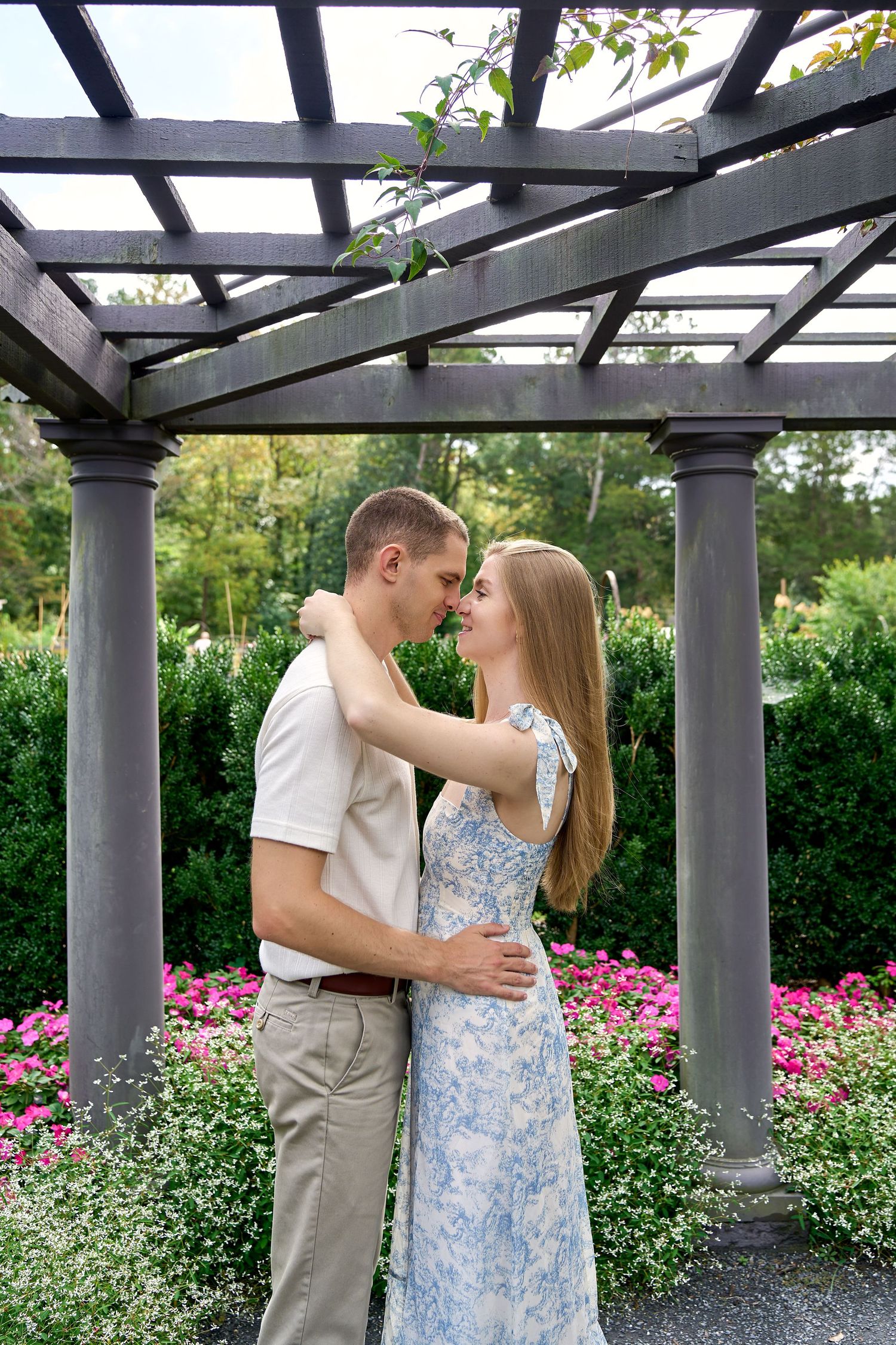 A tender embrace beneath a dark wooden pergola surrounded by colorful garden flowers. North Carolina Wedding Photographer.