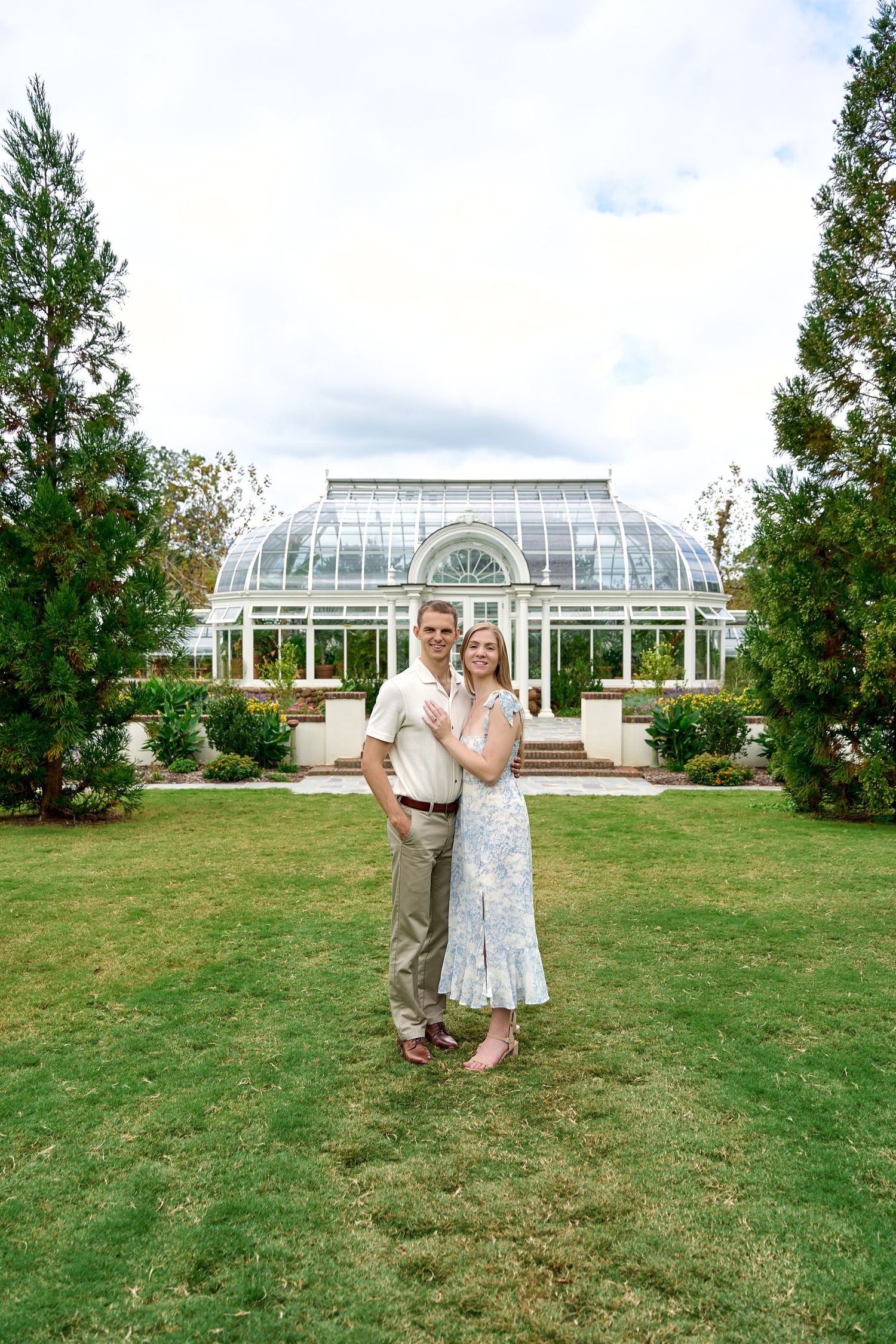 A couple poses in front of a large greenhouse structure on a grassy lawn. North Carolina Wedding Photographer.