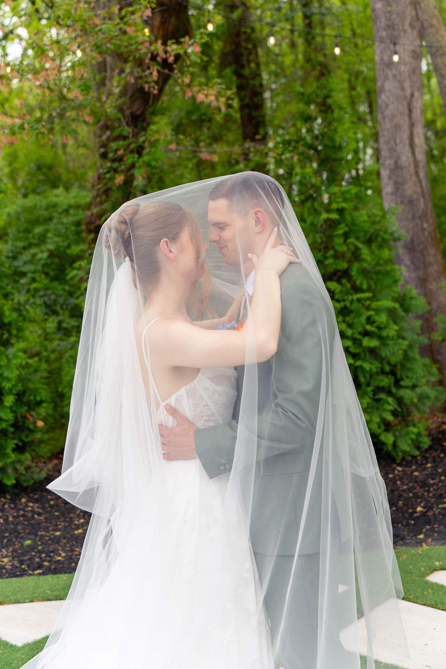 Bride and groom share a romantic kiss hidden beneath brides flowing veil in a lush green garden setting.