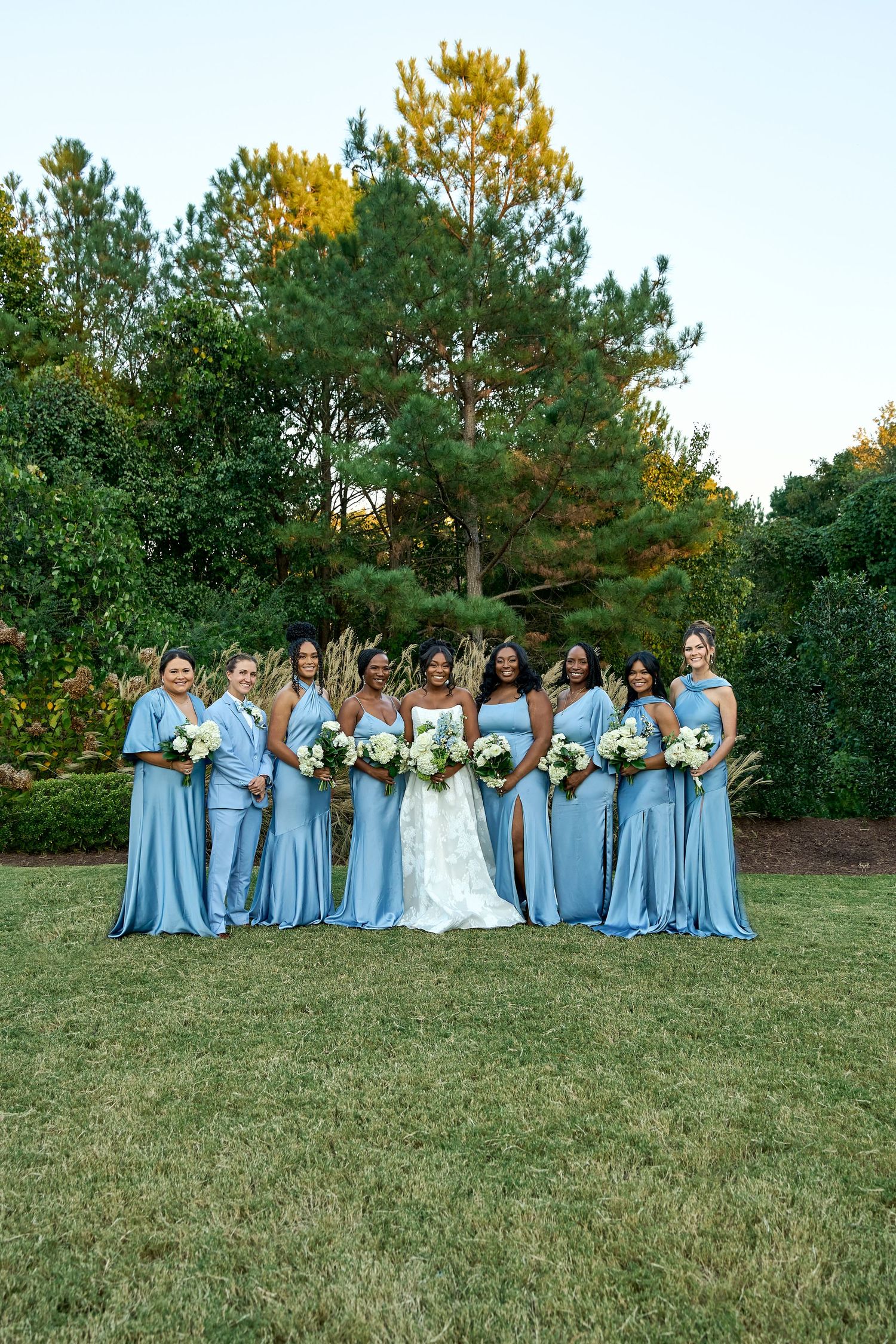 Bride poses with eight bridesmaids wearing matching dusty blue satin gowns in an outdoor garden setting.