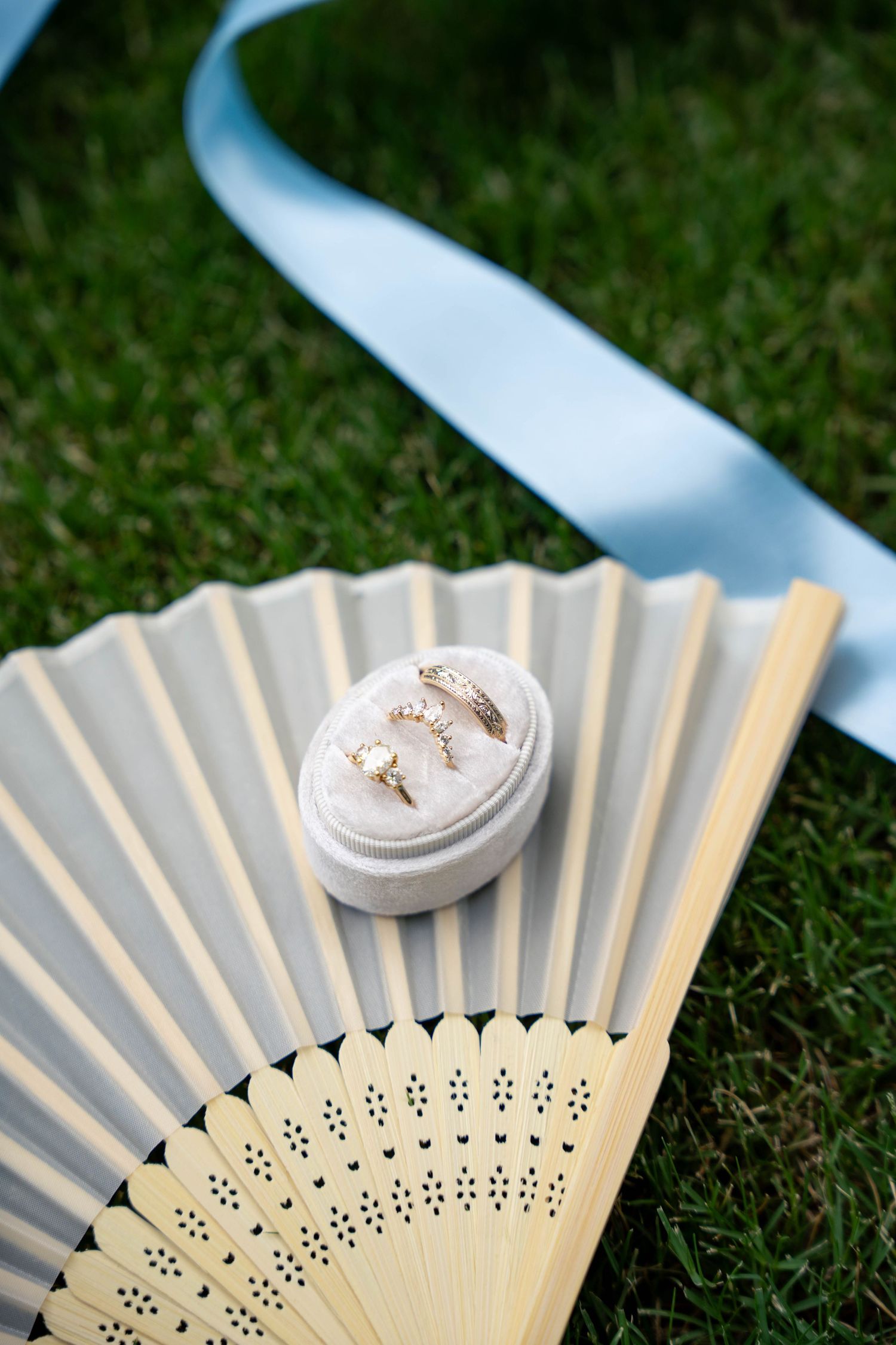 Wedding rings displayed in a velvet box on a wooden fan with a light blue ribbon on green grass.