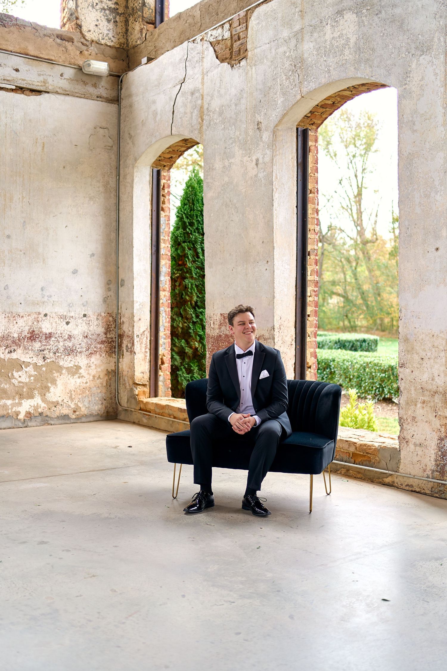 Groom in black tuxedo sits in velvet chair inside rustic brick ruins venue. North Carolina Wedding Photographer.