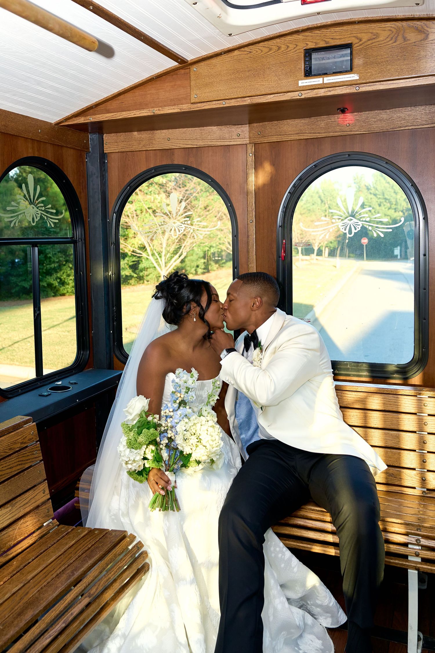 Newlywed couple sharing a kiss inside a vintage wooden trolley, bride holding blue and white bouquet.