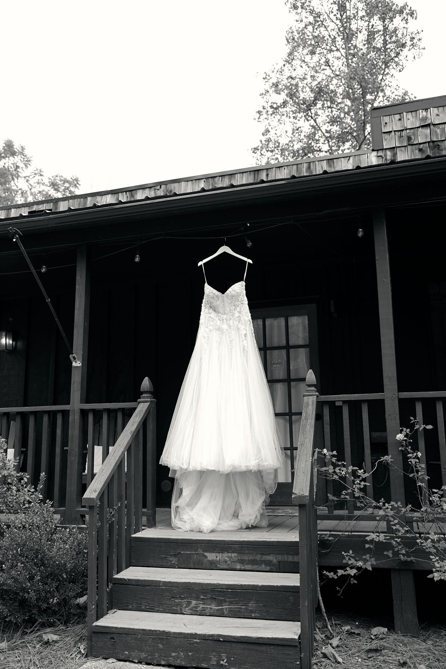 White wedding gown hanging on a hanger on the porch of a rustic dark wood building, black and white photo.