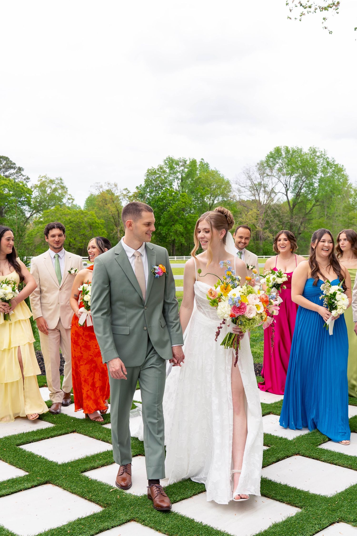 Bride and groom walk hand-in-hand down the aisle flanked by colorful bridesmaids and groomsmen at outdoor wedding.