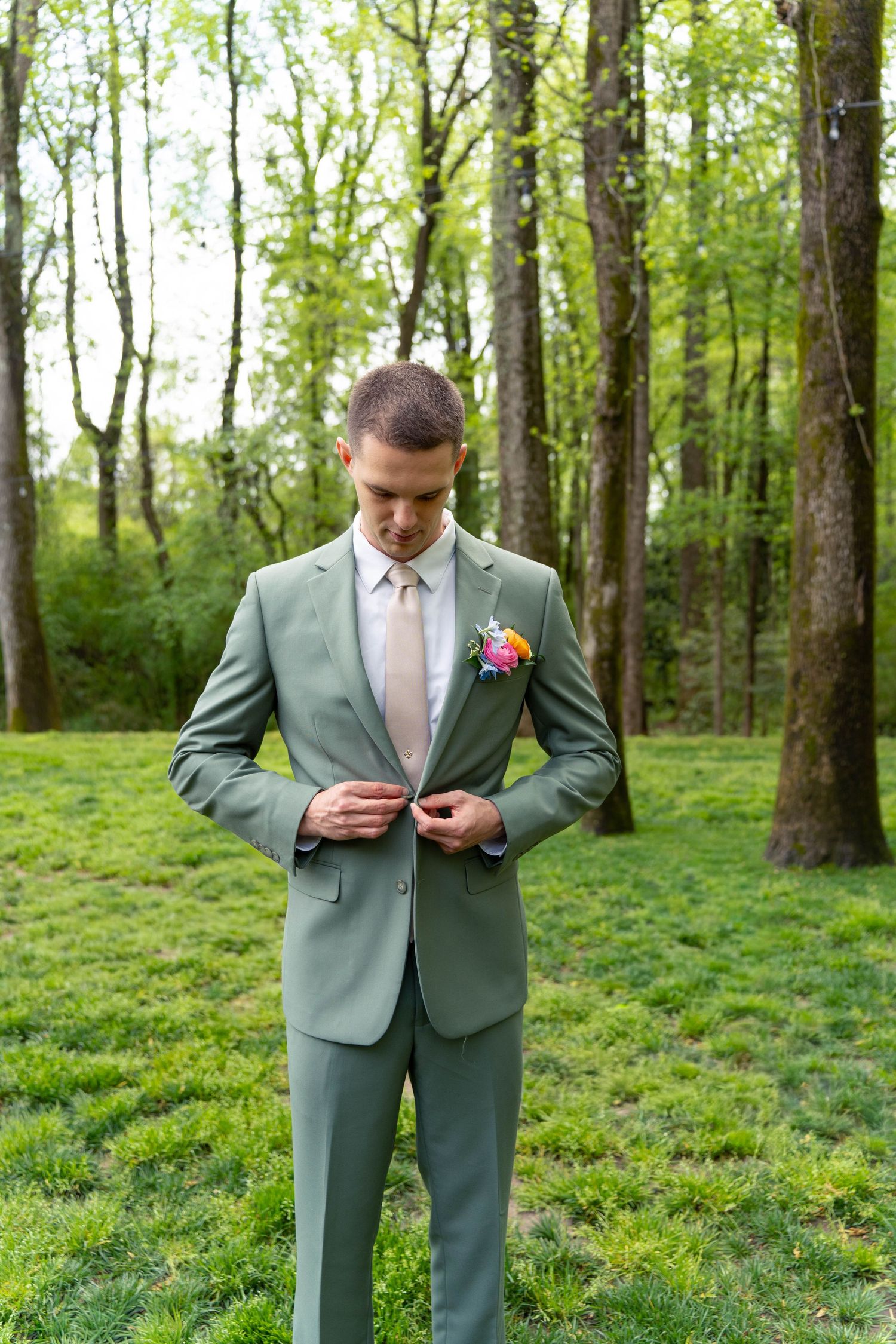 Groom in sage green suit buttons his jacket while standing in a lush green wooded outdoor setting.
