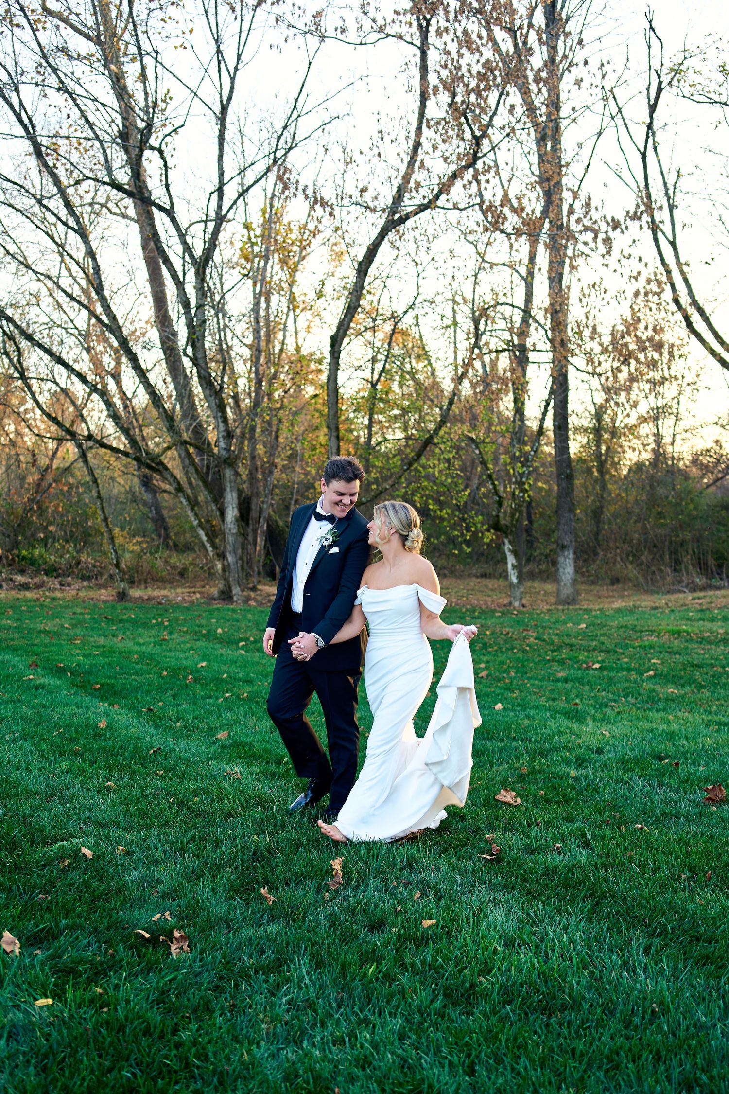 A couple in wedding attire walks together on a green lawn with autumn trees in the background at sunset.