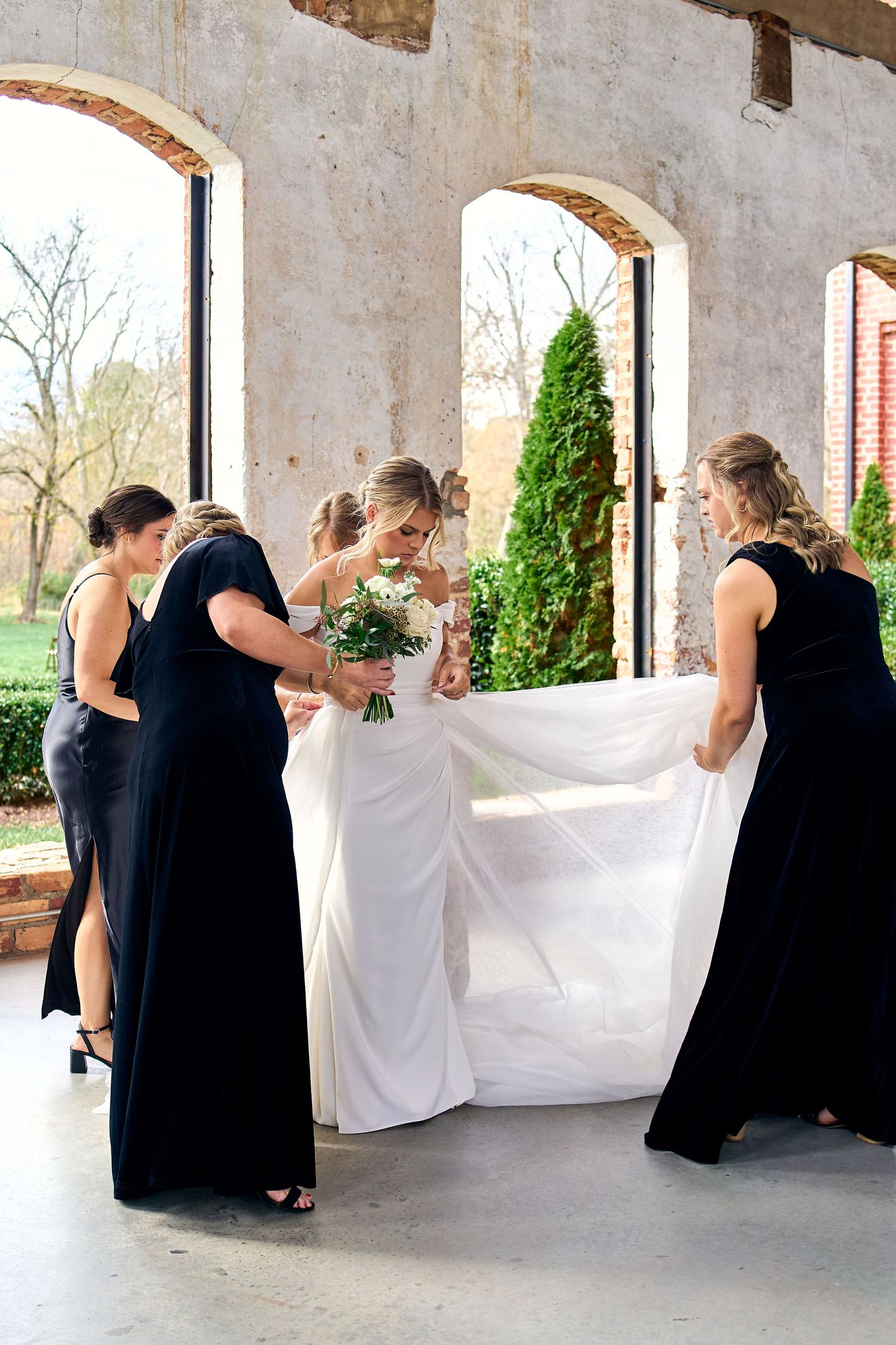 Wedding party members in black dresses help adjust a flowing white bridal gown in an elegant columned venue.