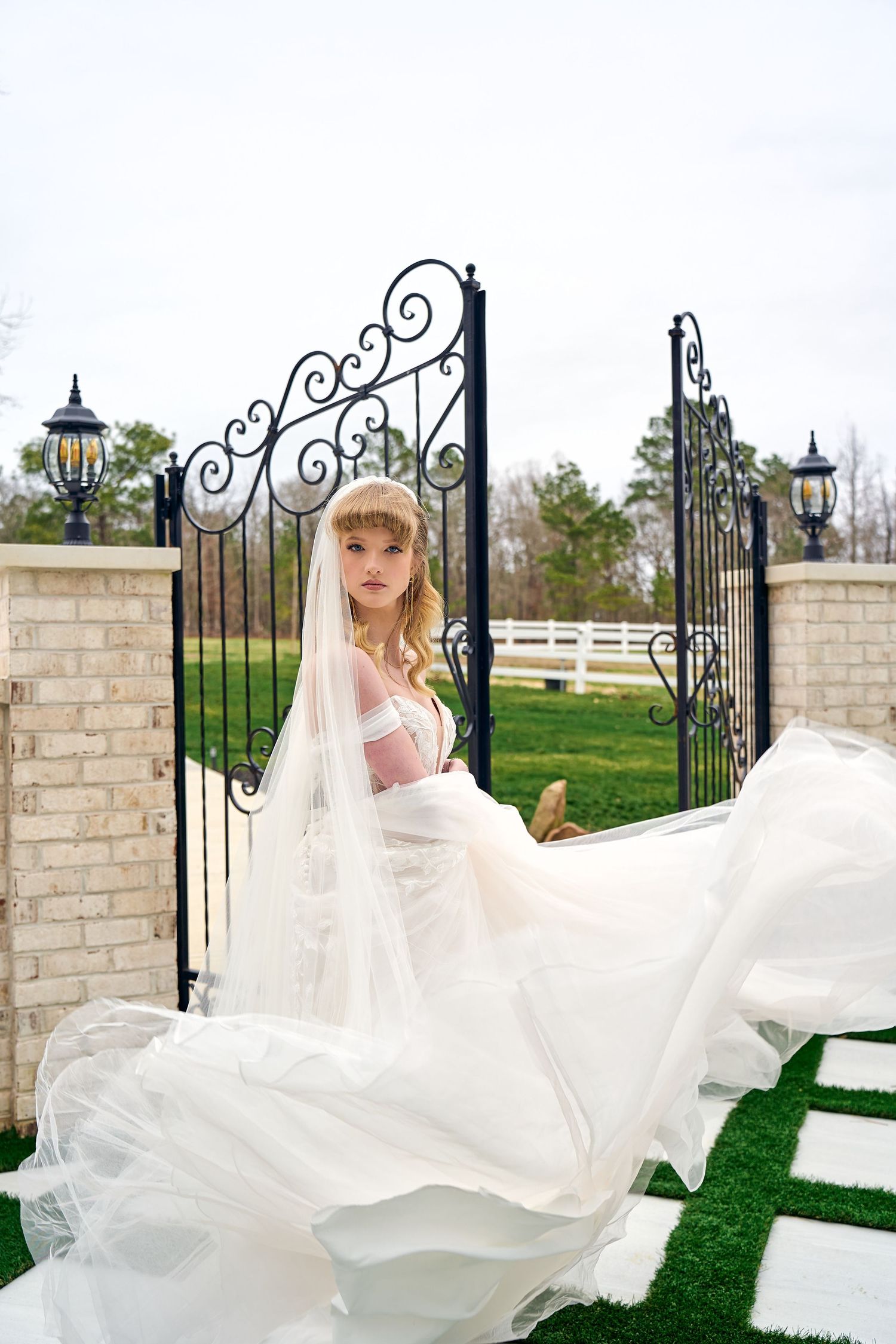 An elegant flowing white wedding dress billows in the breeze by ornate wrought iron gates and brick columns.
