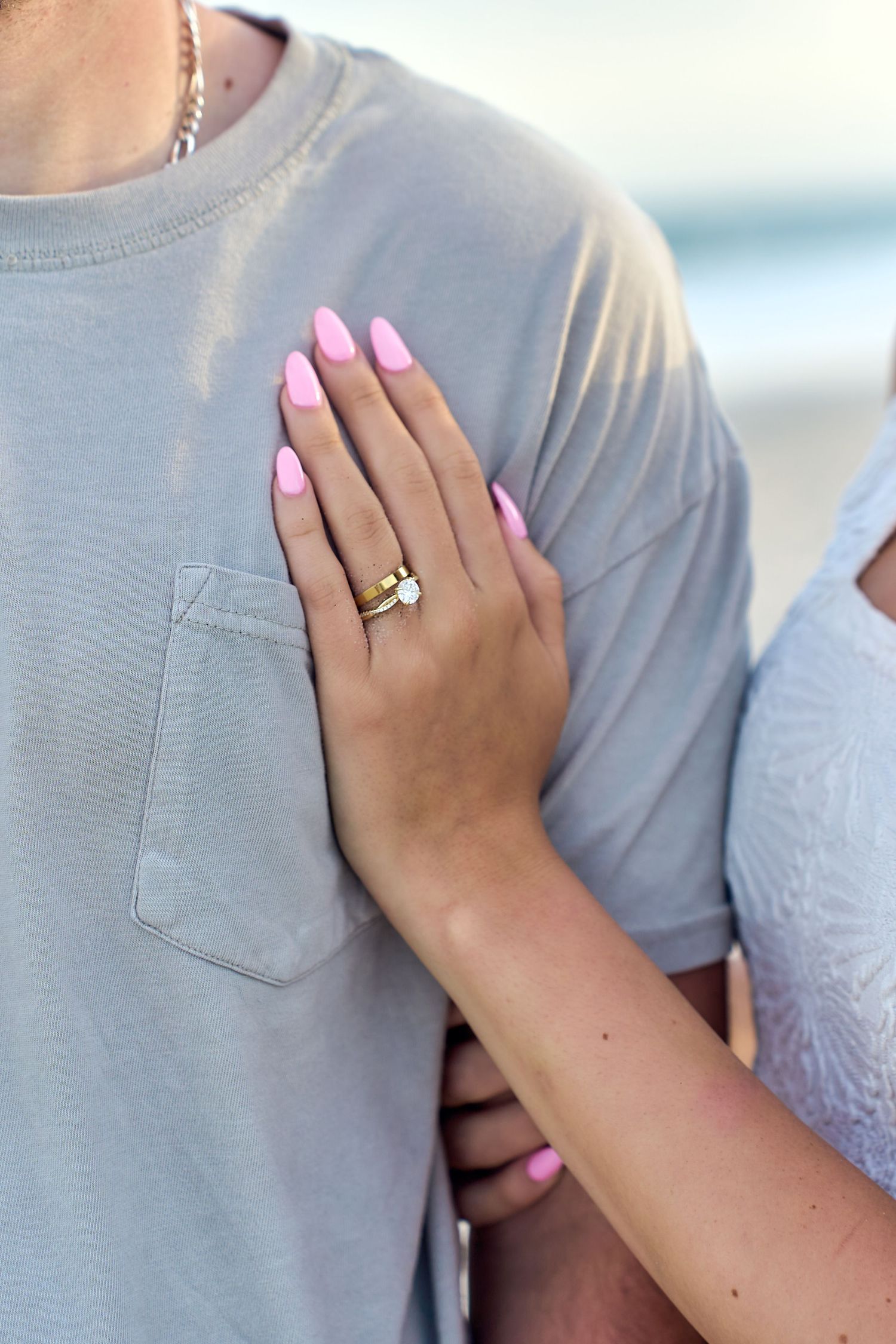 Close up of a hand with pink manicured nails and engagement ring resting against a gray t-shirt.
