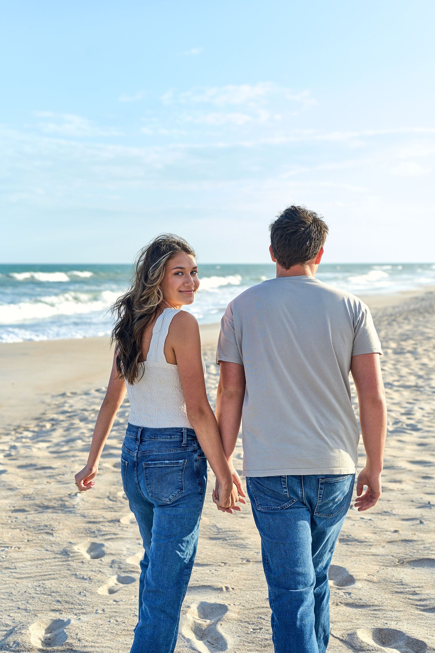 A couple holding hands while walking together on a sunny beach with waves in the background.