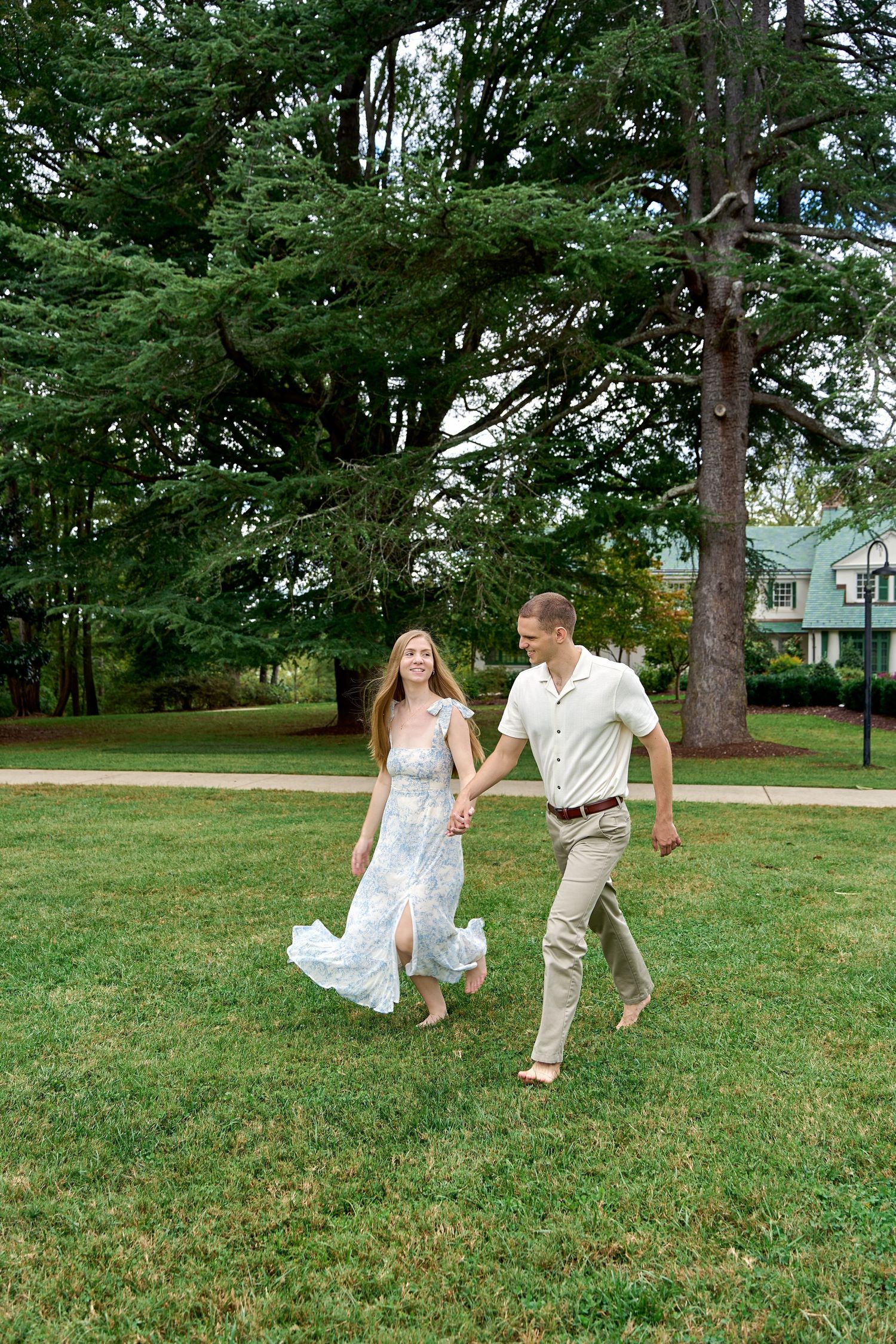 A couple runs playfully through a grassy field surrounded by trees on a sunny day. North Carolina Wedding Photographer.