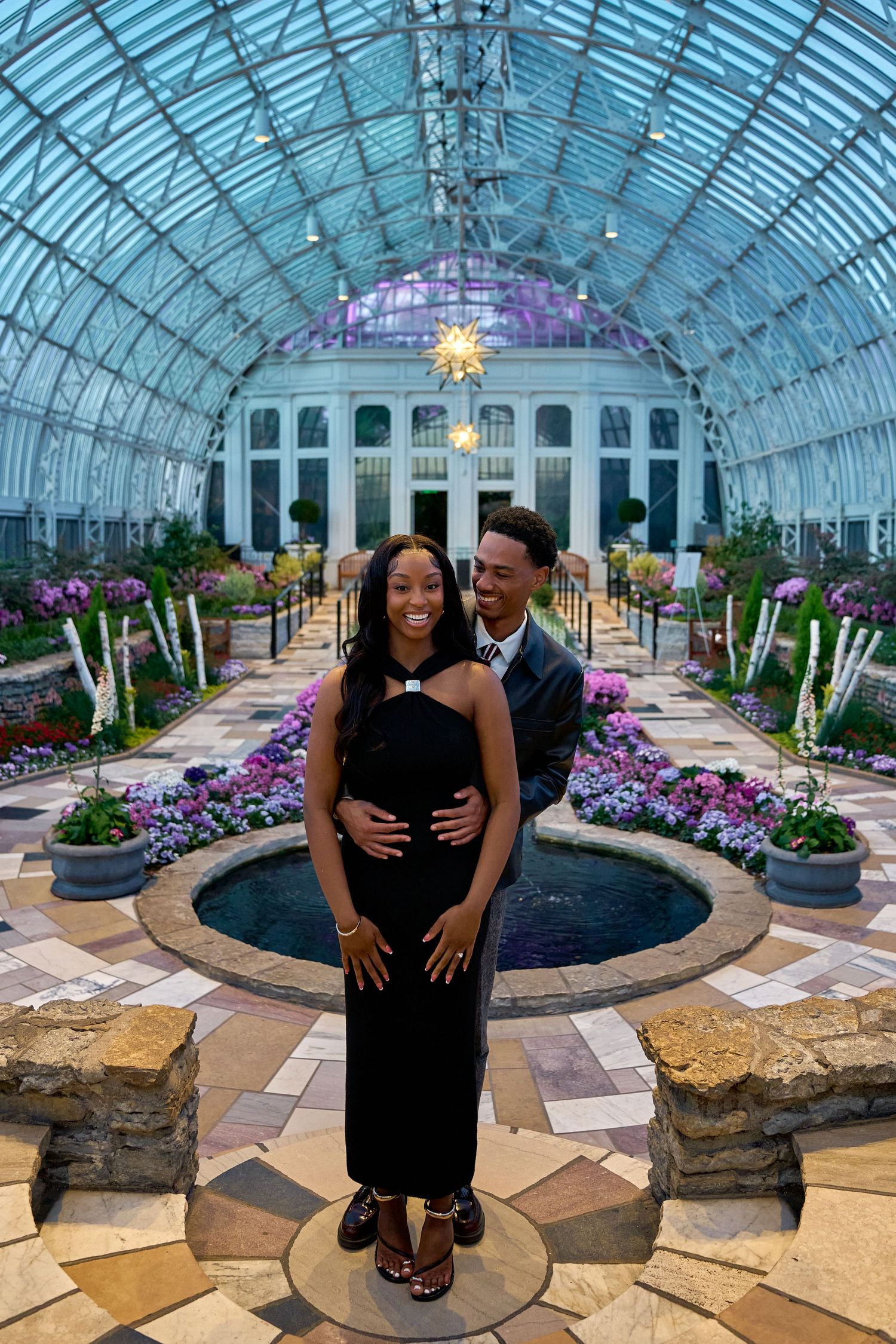 Couple posing together inside a stunning glass conservatory with a fountain and colorful floral displays.