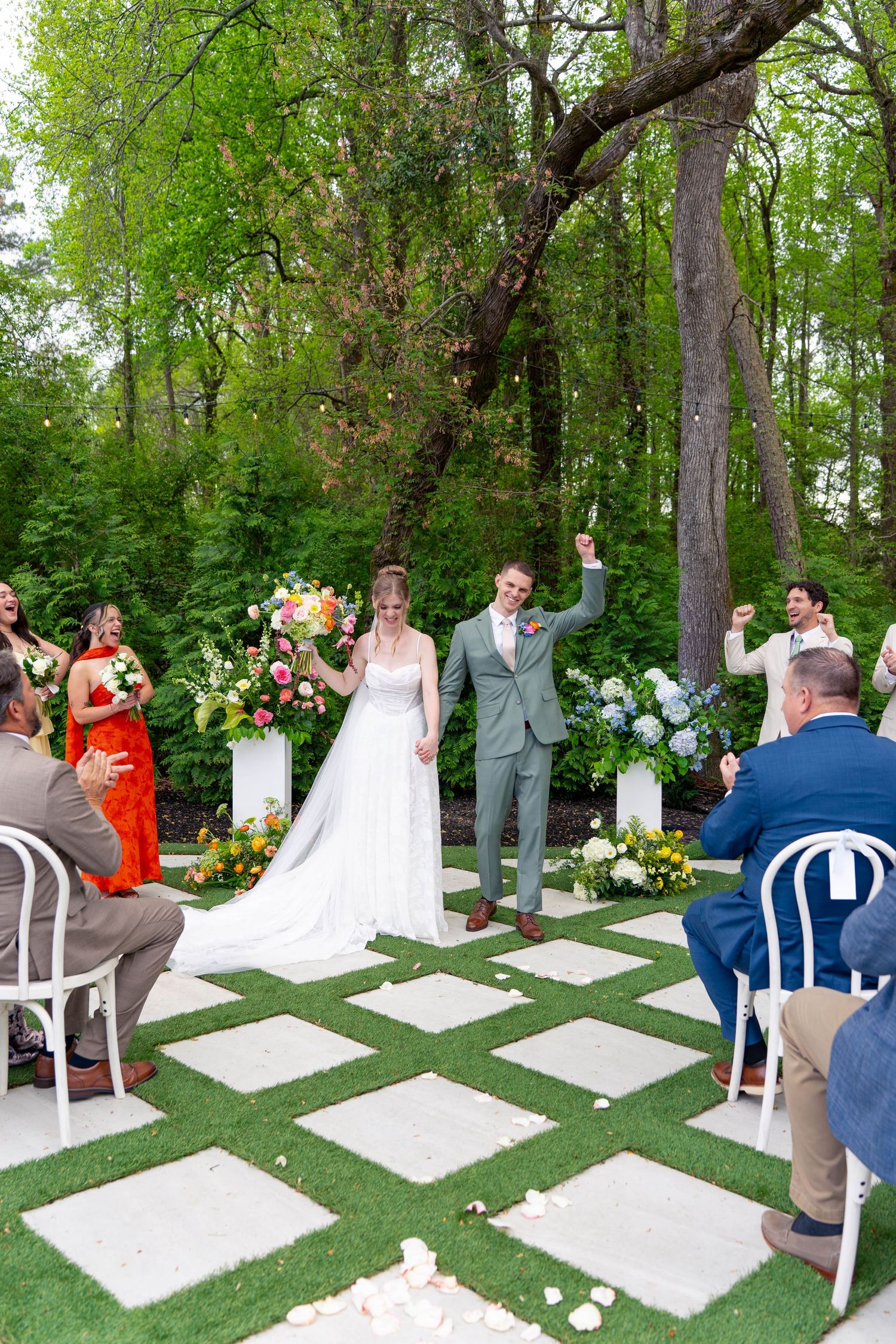 Jubilant bride and groom raise hands walking back down checkered aisle after outdoor wedding ceremony.