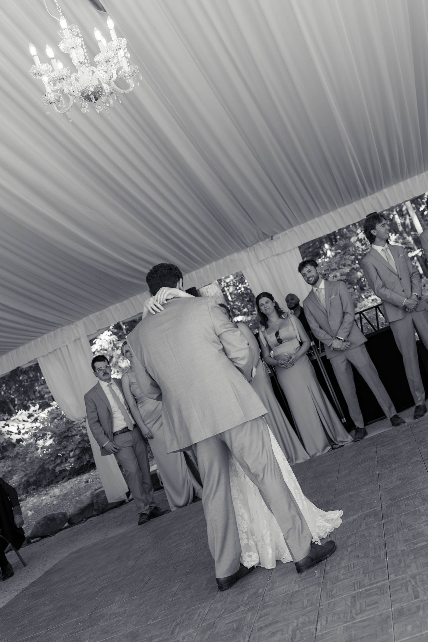 Black and white photo of bride and groom first dance with wedding party watching under tent.