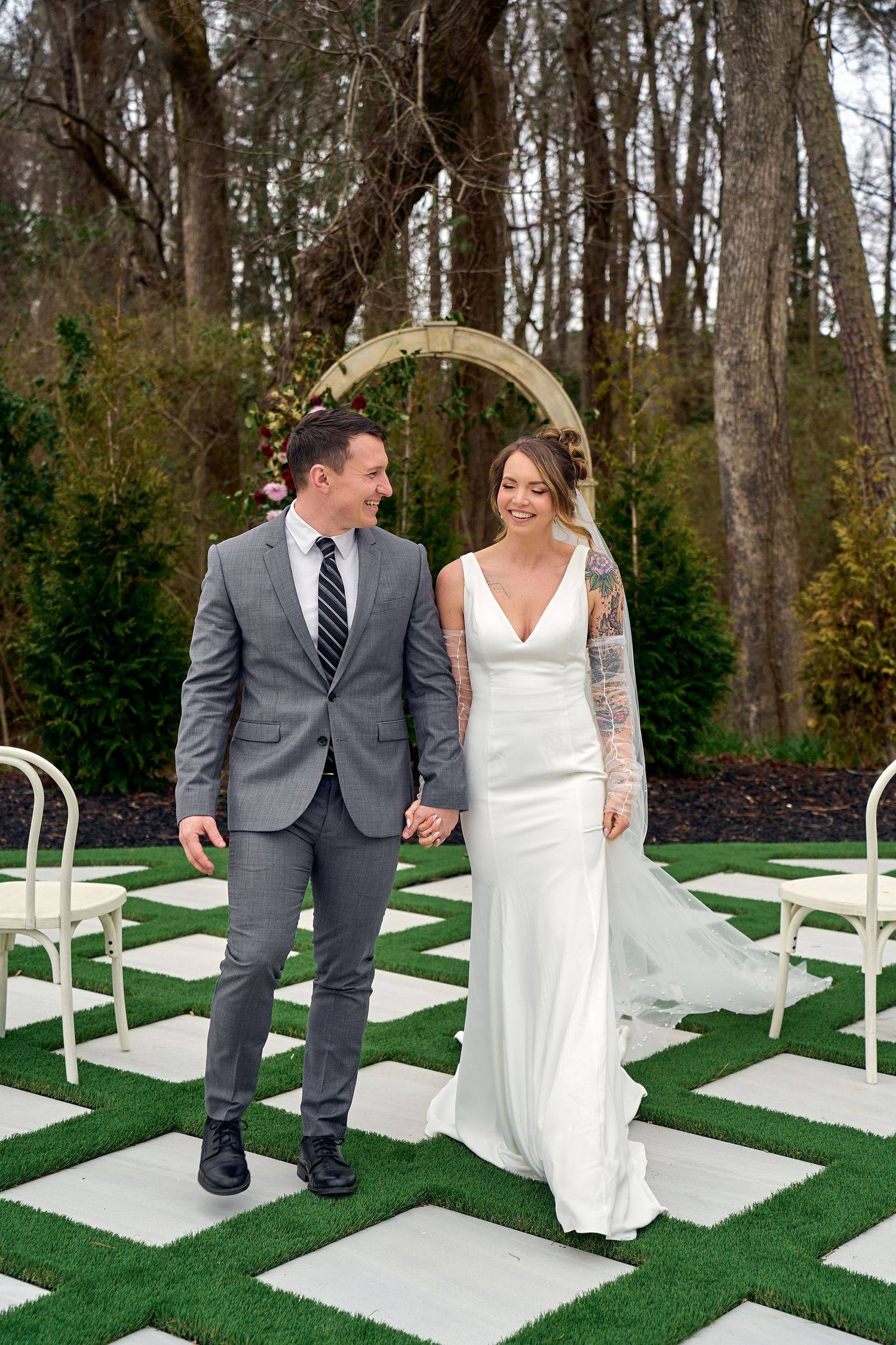 A couple shares a joyful moment on a checkerboard grass and stone ceremony area under a golden arch in a wooded setting.