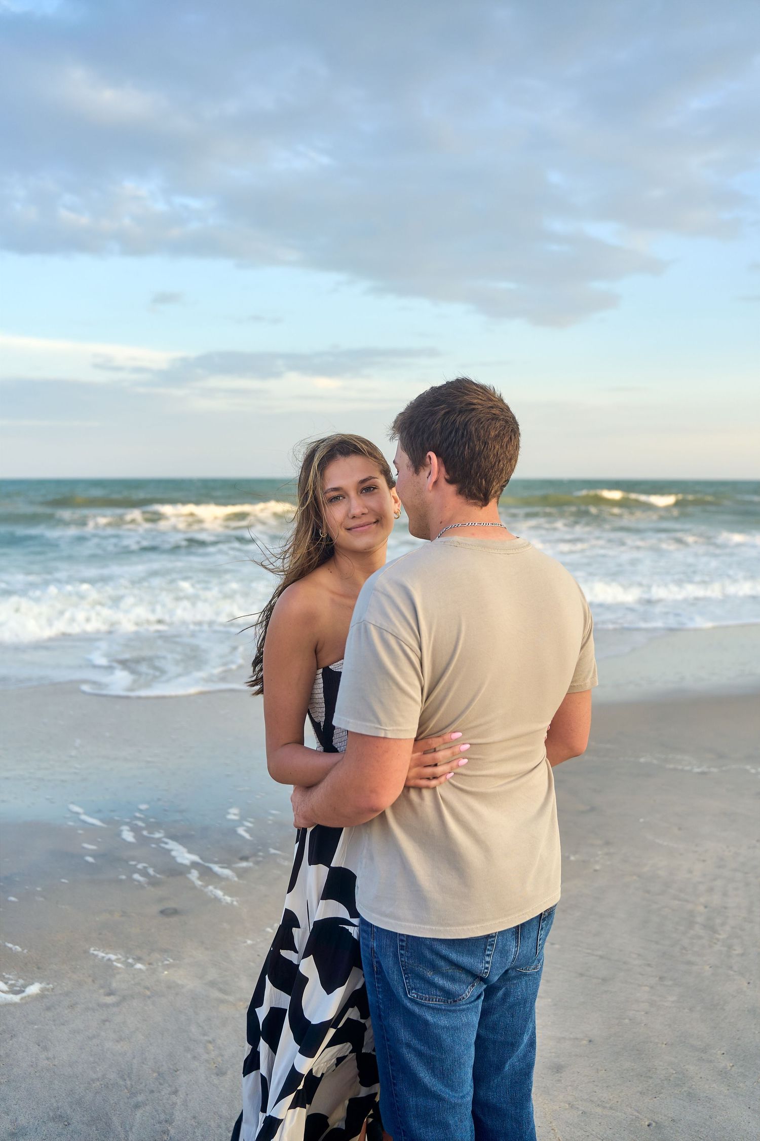 A romantic couple embraces while standing on a beach at sunset with waves crashing in the background.
