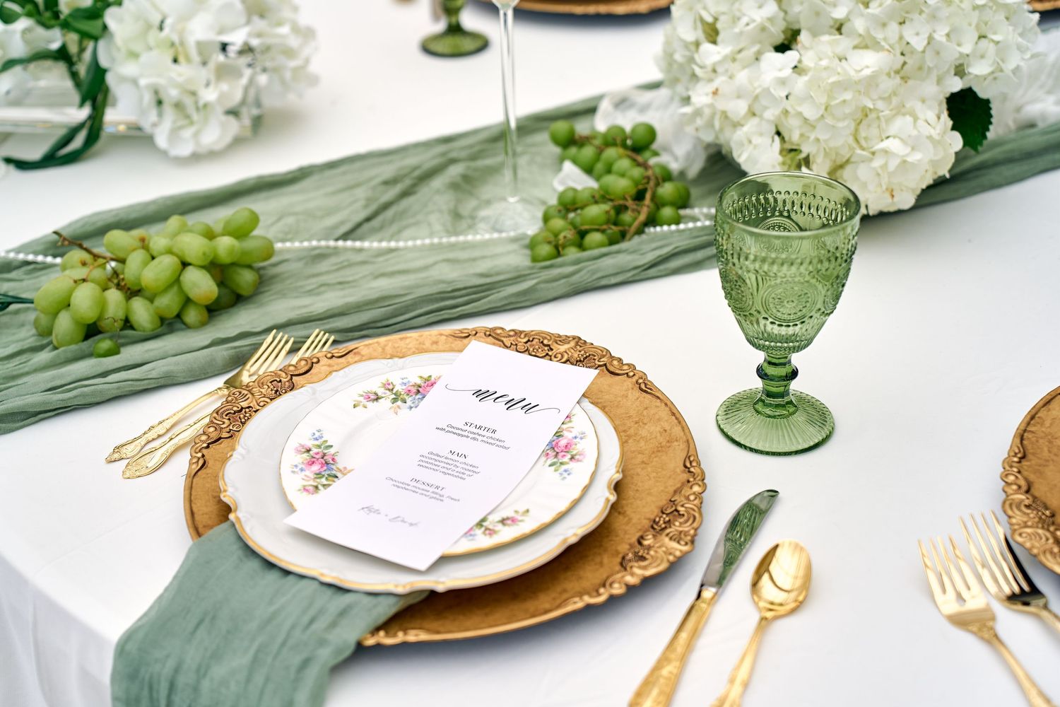 Elegant table setting with gold charger plates, vintage green glassware, white flowers and fresh grapes as decor.