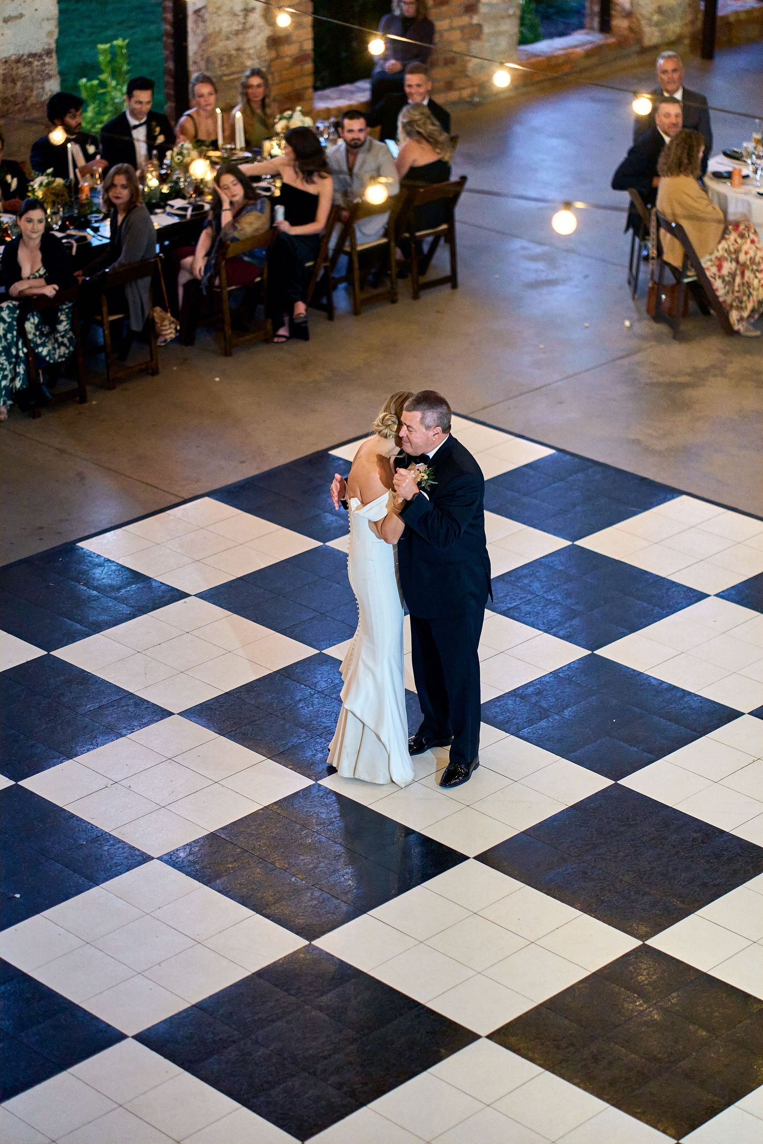 A couple shares their first dance on a black and white checkered dance floor while guests watch from tables in a rustic venue.