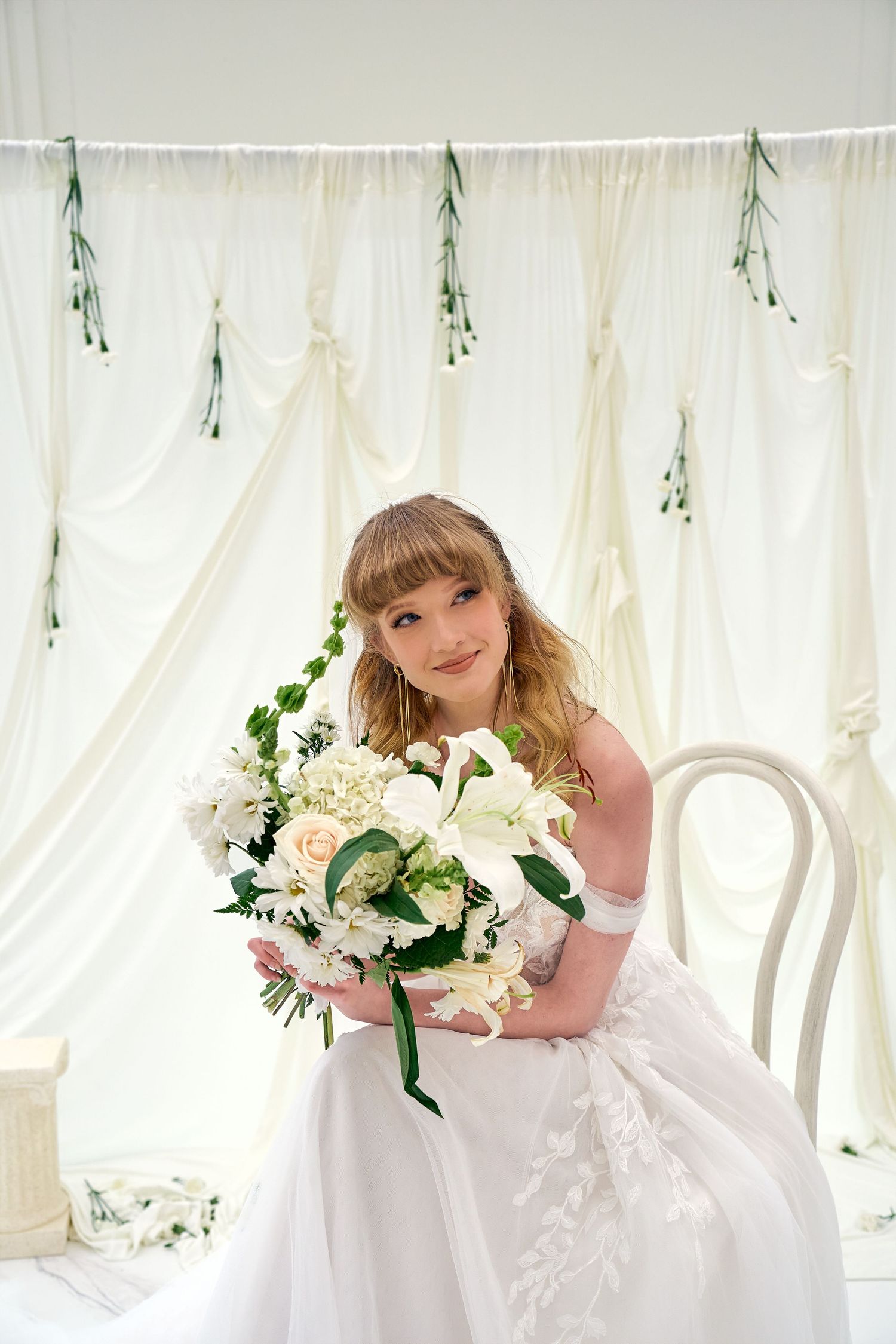 A bride in an off-shoulder white wedding dress holds a lush bouquet of white flowers against a draped fabric backdrop.