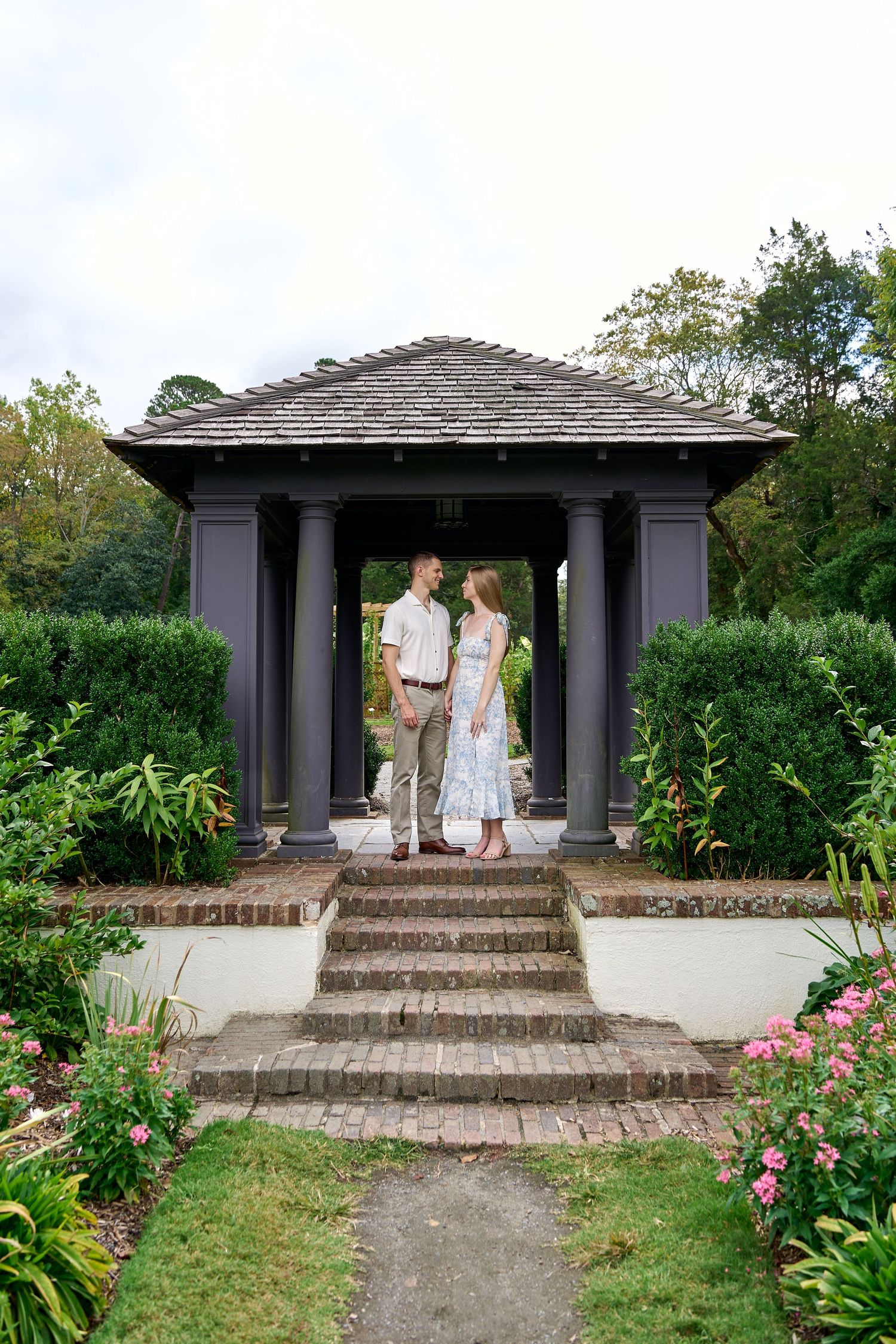 Couple holding hands and facing each other under an elegant dark gazebo surrounded by garden greenery.