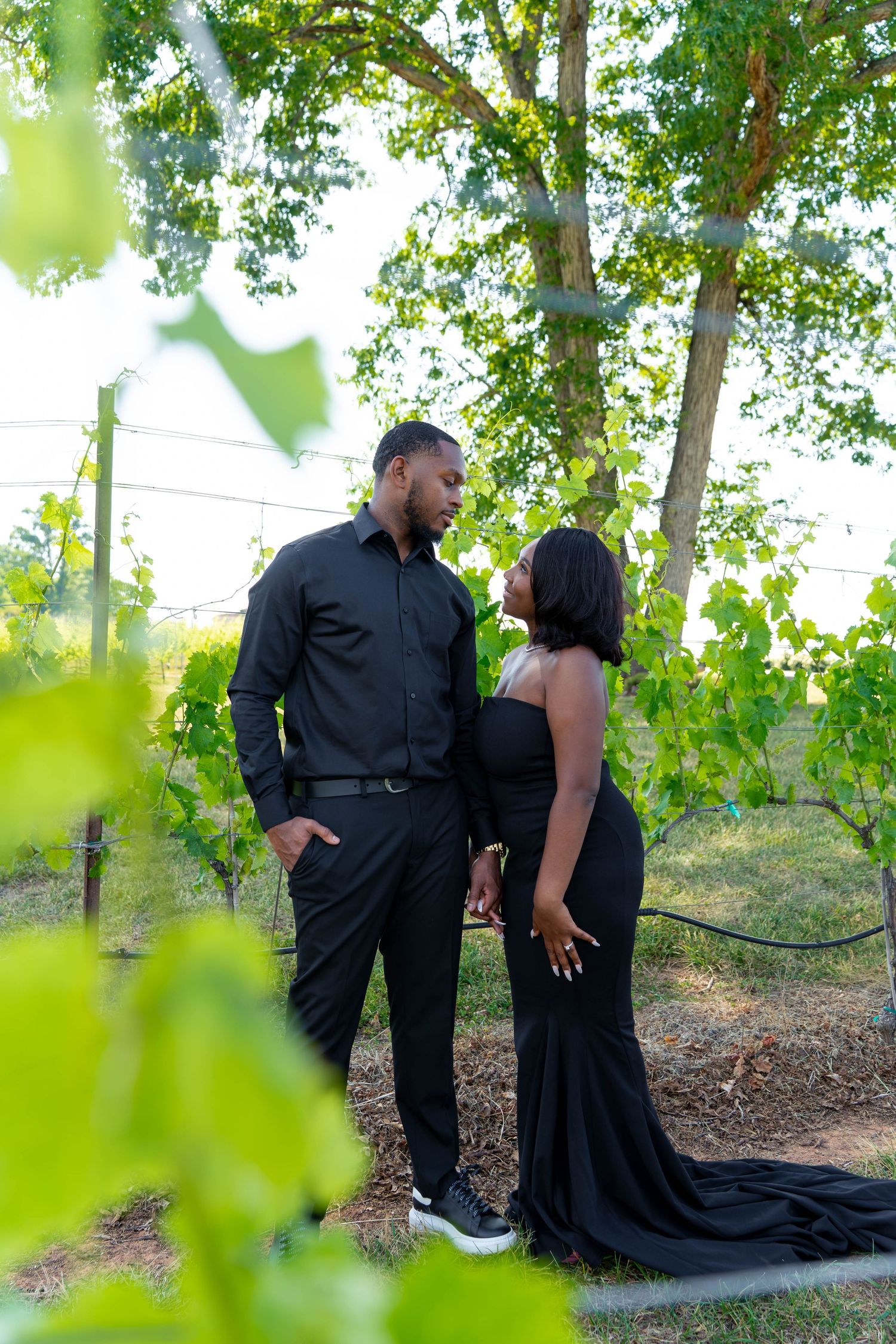 Couple dressed in black hold hands and gaze at each other among vineyard grapevines.