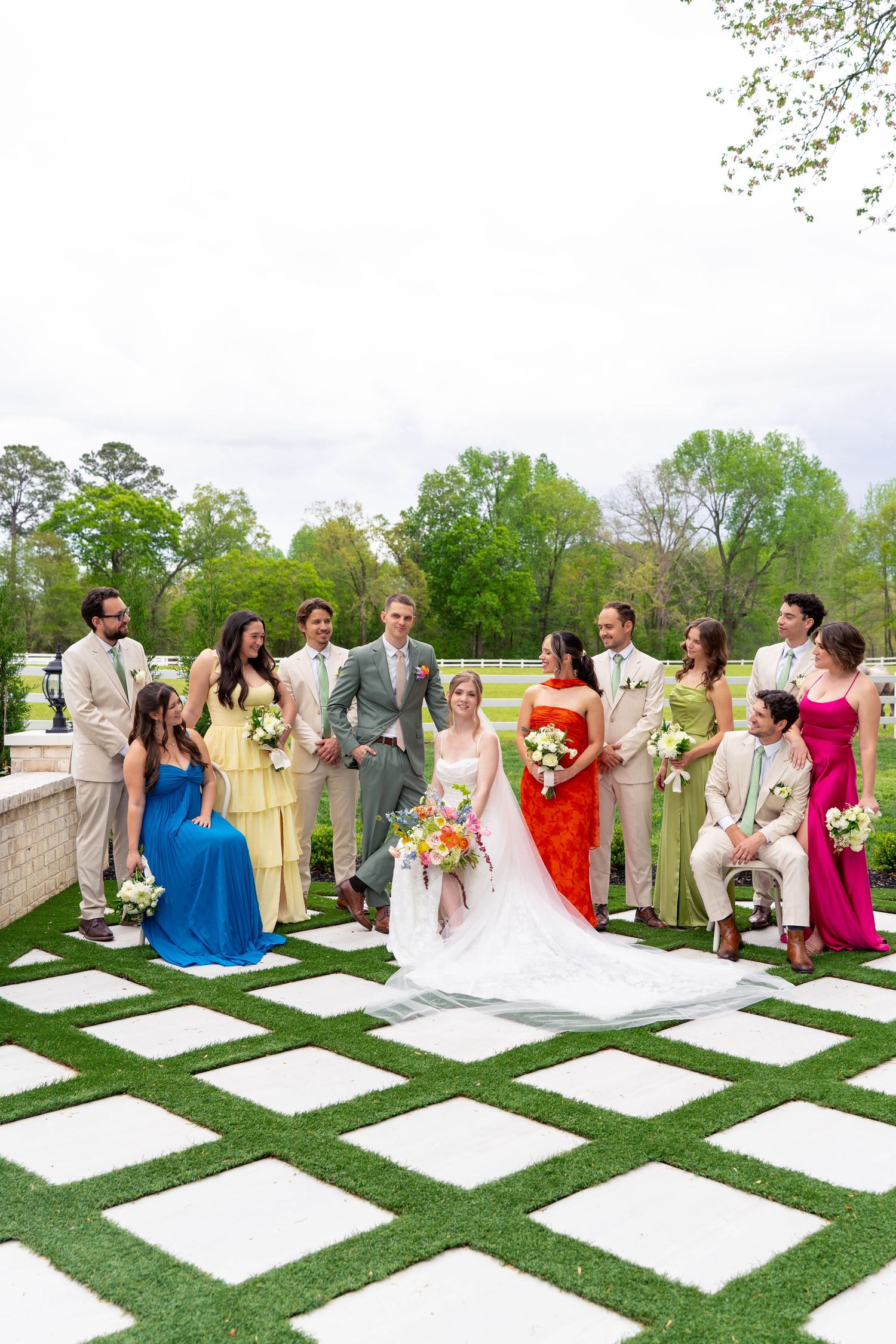 Wedding party in colorful attire posing on checkered grass and stone patio at outdoor venue.
