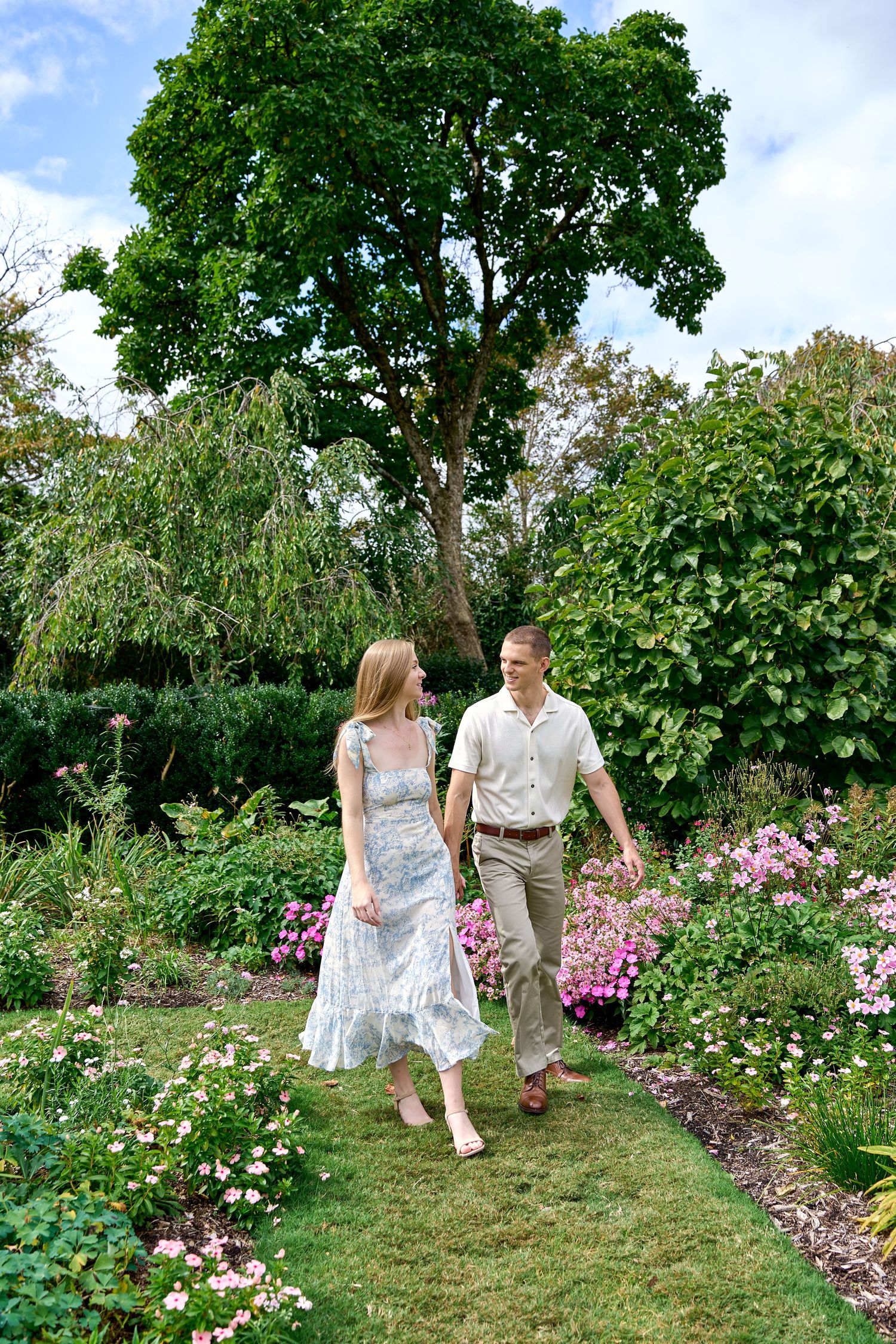 Couple walking hand-in-hand along a lush garden path lined with pink flowers and towering green trees.