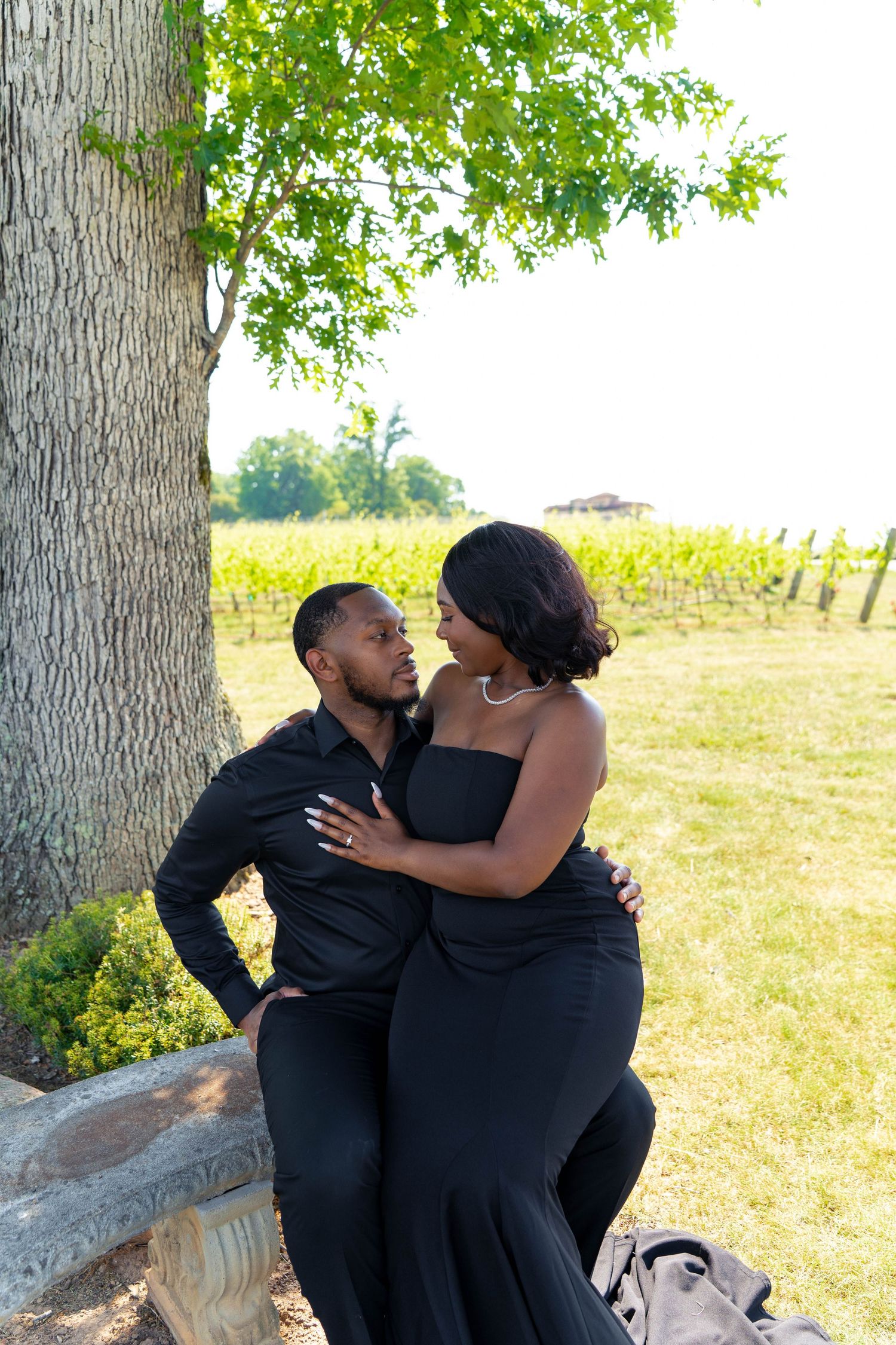 Couple in matching black outfits share a romantic moment by a tree at a vineyard.