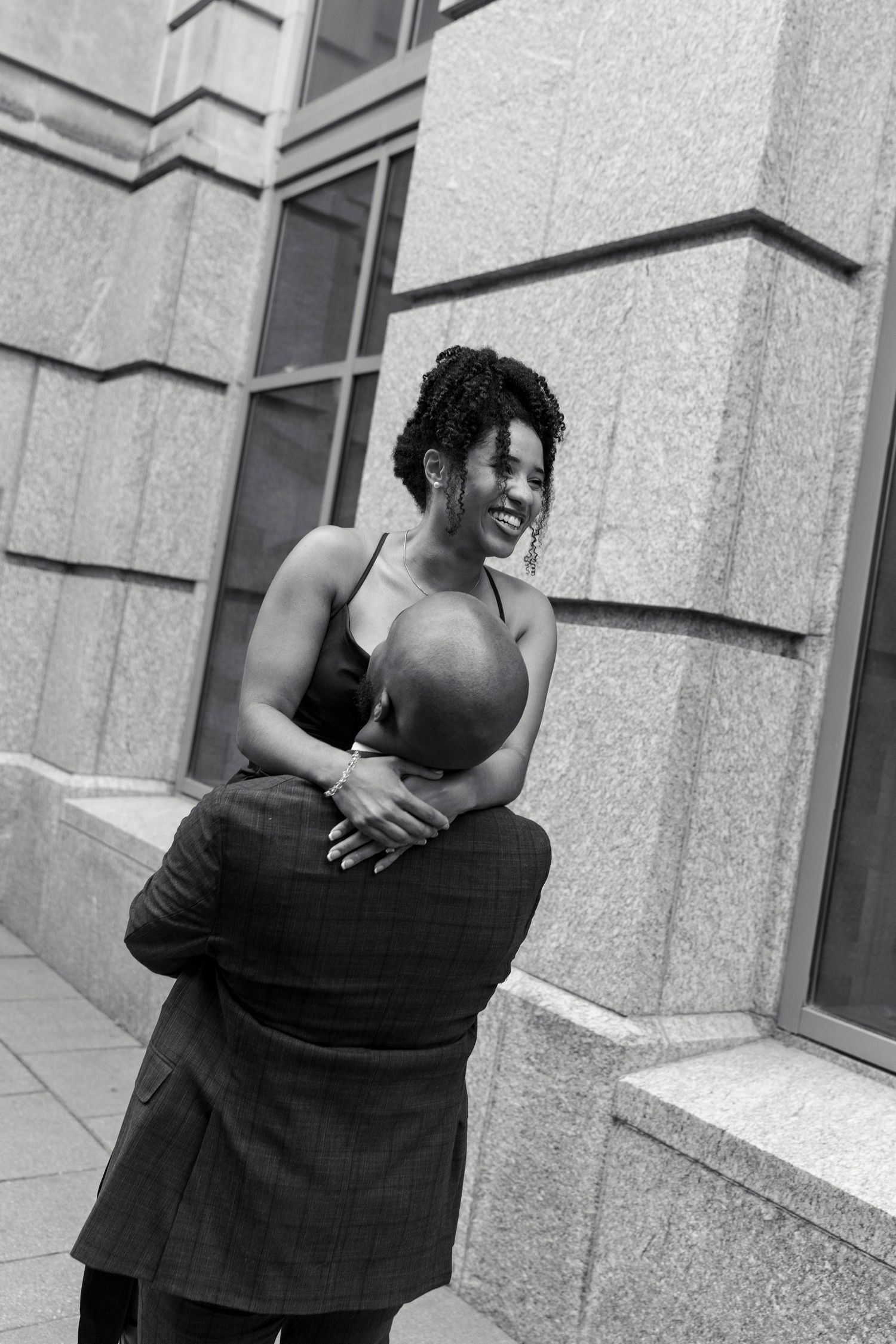 Black and white photo of a man lifting a laughing woman in a satin dress beside a stone wall.
