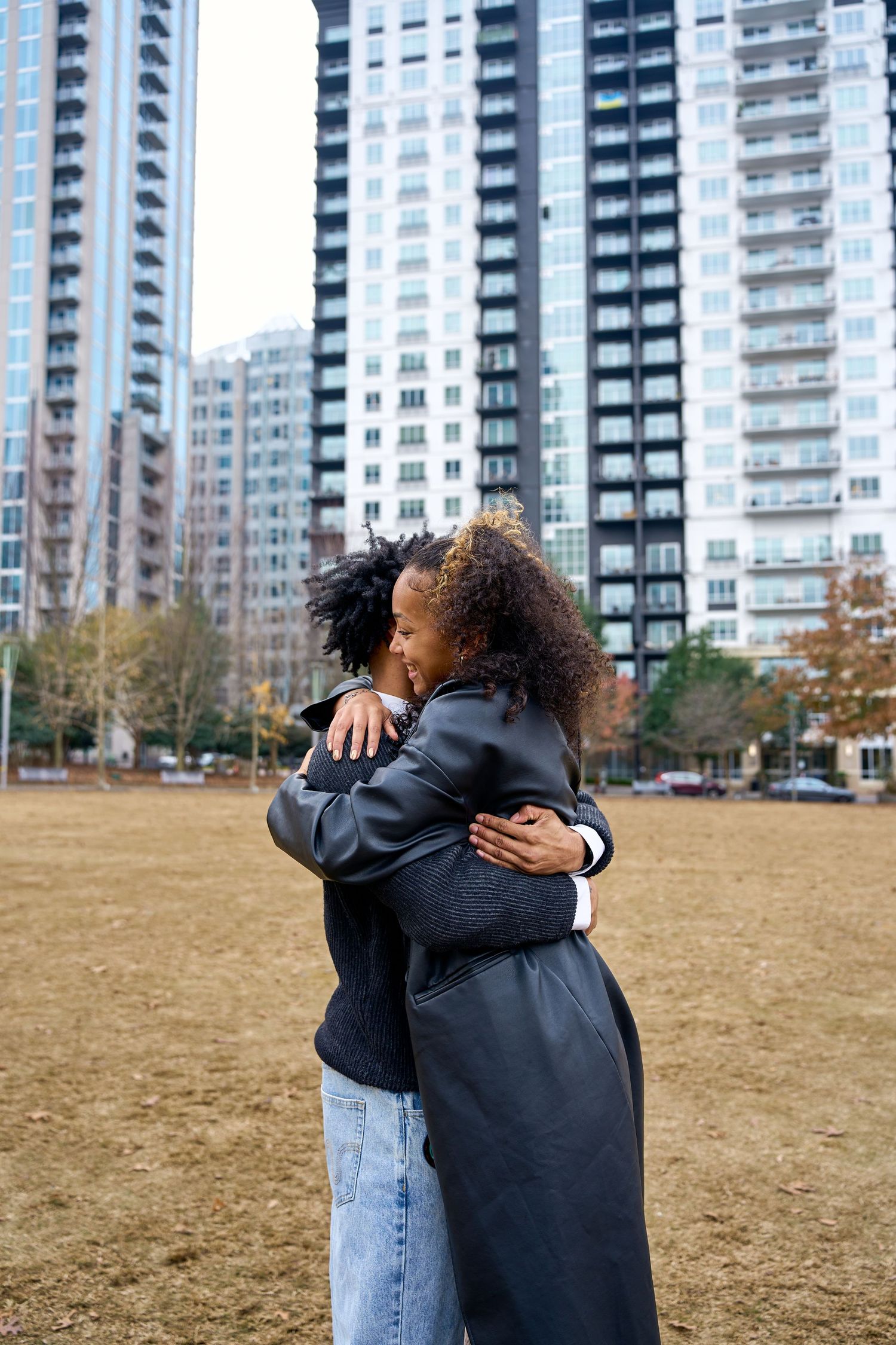 Two people share an emotional embrace in a park with tall apartment buildings visible in the background. North Carolina Wedding Photographer