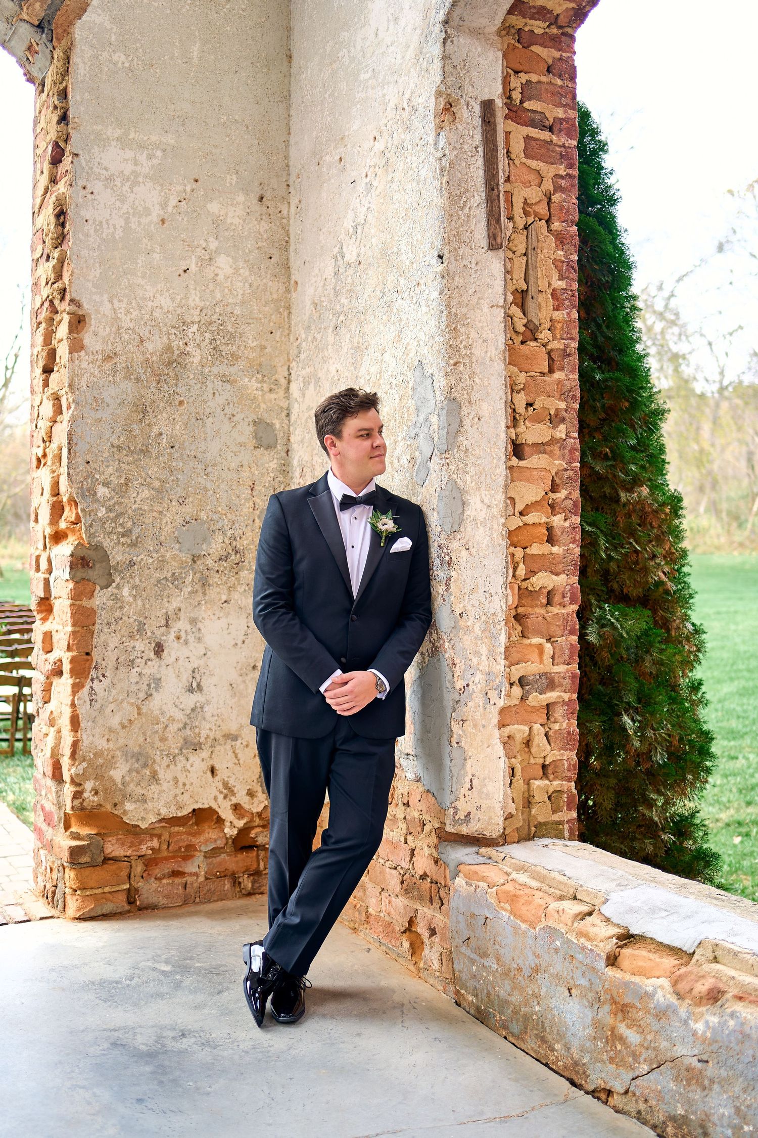 A groom in a black tuxedo and bow tie leans against an aged brick column at a rustic outdoor venue.