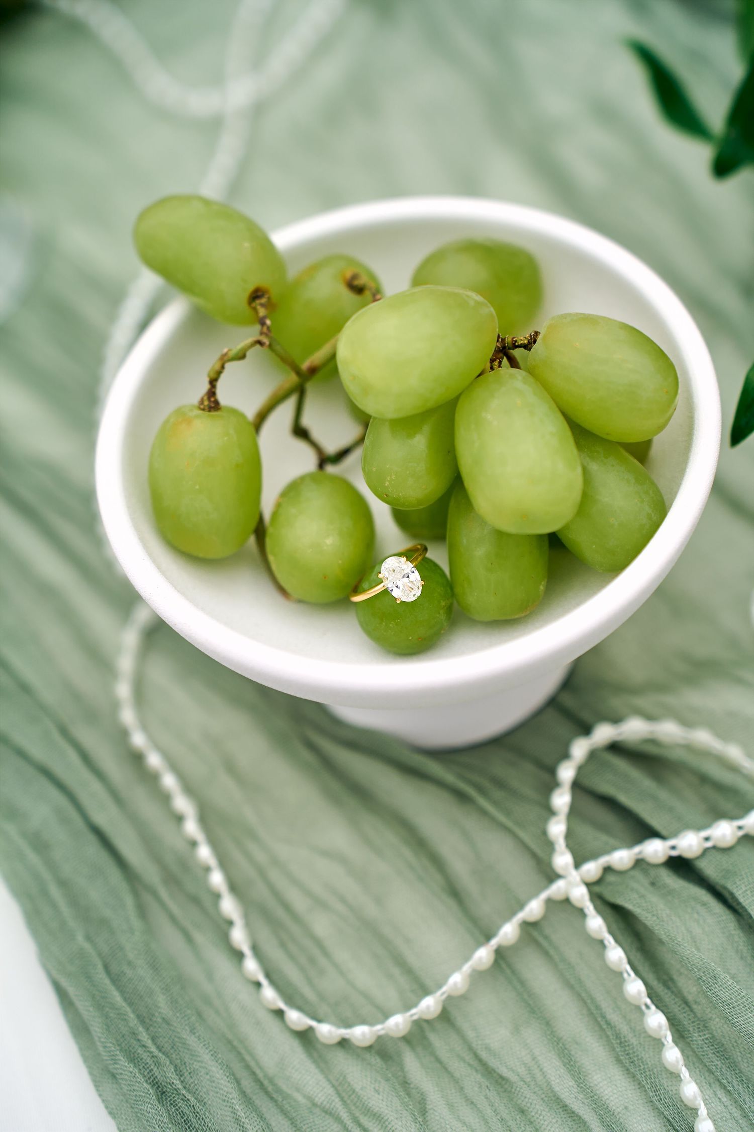 Fresh green grapes in a white ceramic bowl with a delicate pearl necklace on a mint green surface.