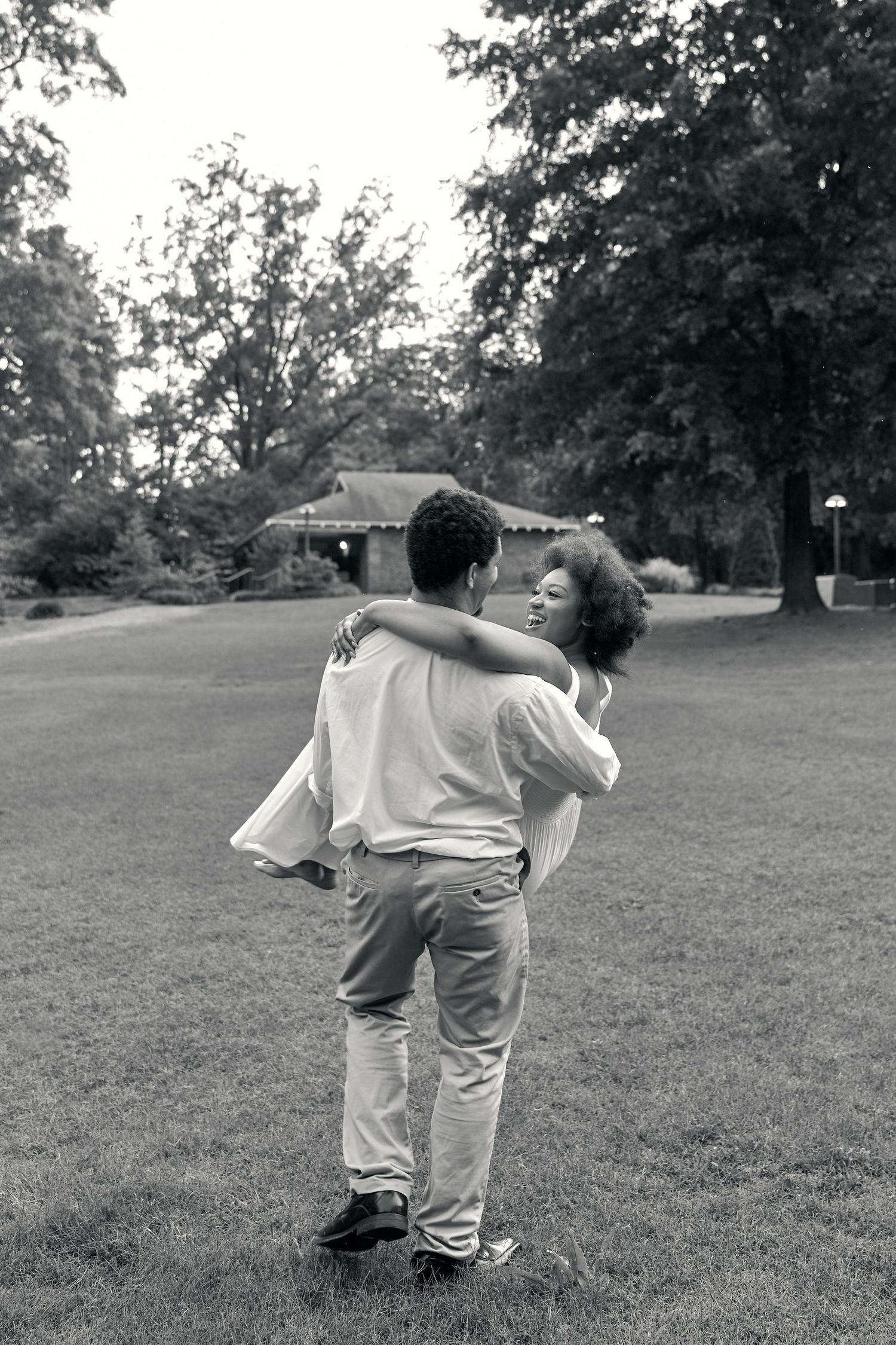 A playful couple enjoys a piggyback ride moment in a park setting with trees in the background.