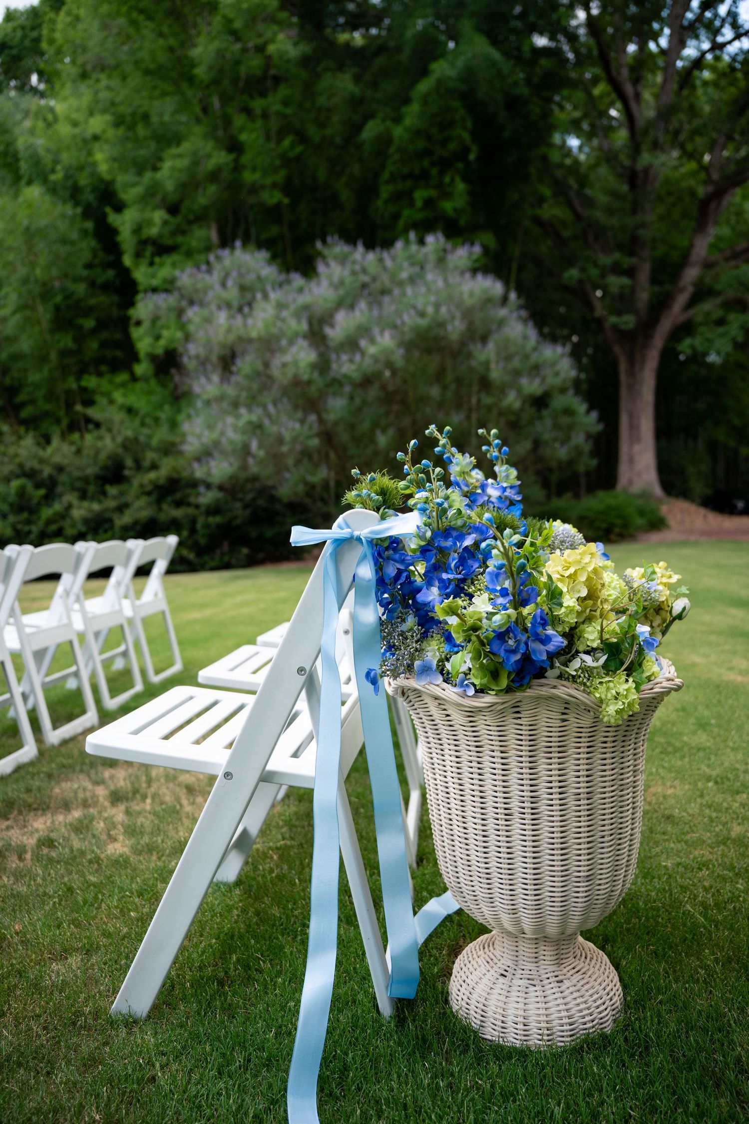 Elegant white wicker urn filled with blue and green flowers with light blue ribbon decorates outdoor wedding aisle.