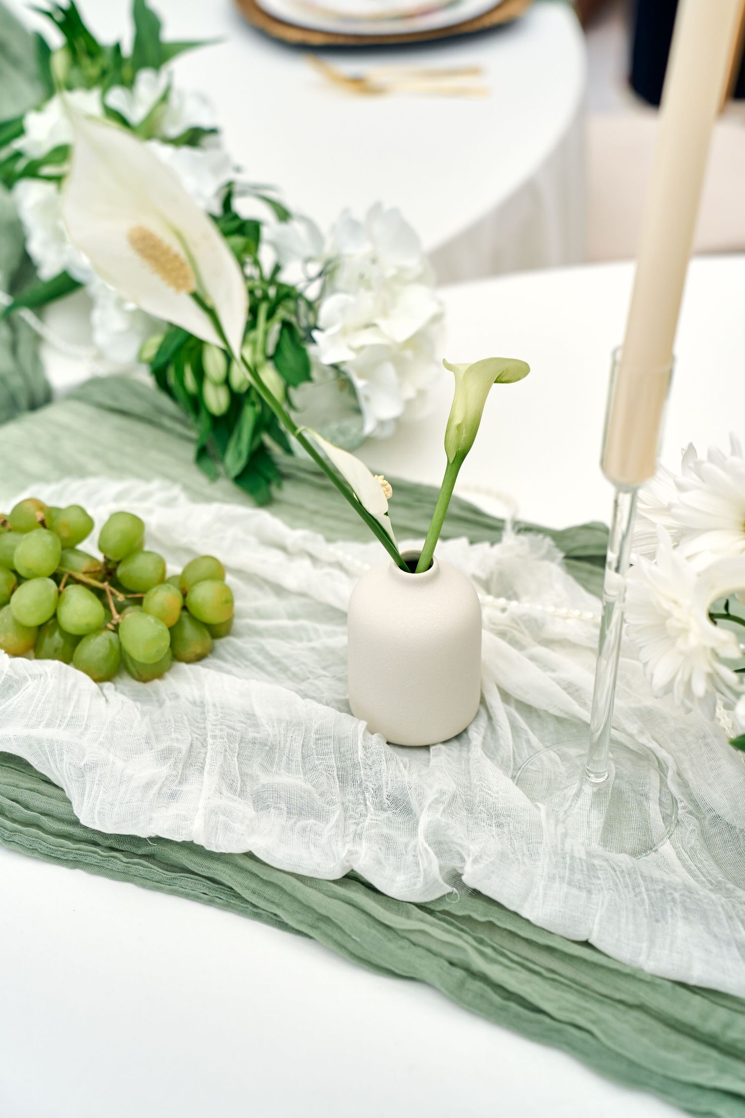 Elegant table setting with green grapes, white flowers and gauzy sage green fabric on a white surface.