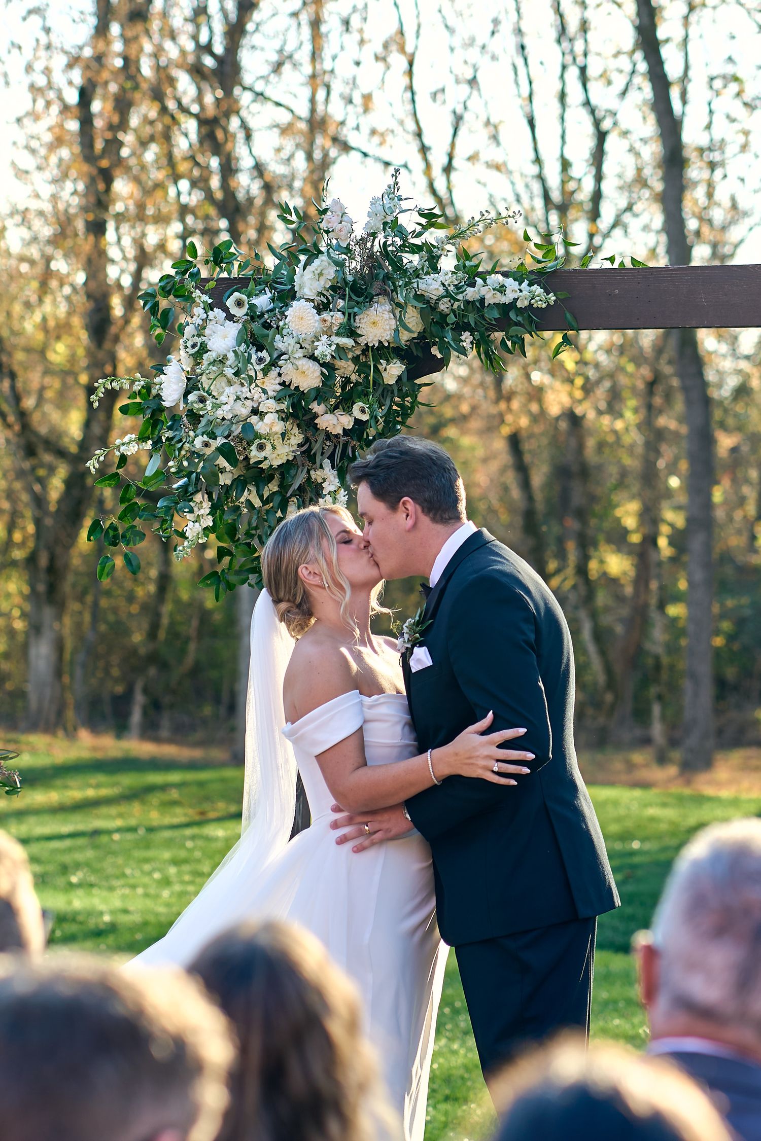 A bride and groom share their first kiss beneath a floral arch during an outdoor autumn wedding ceremony.