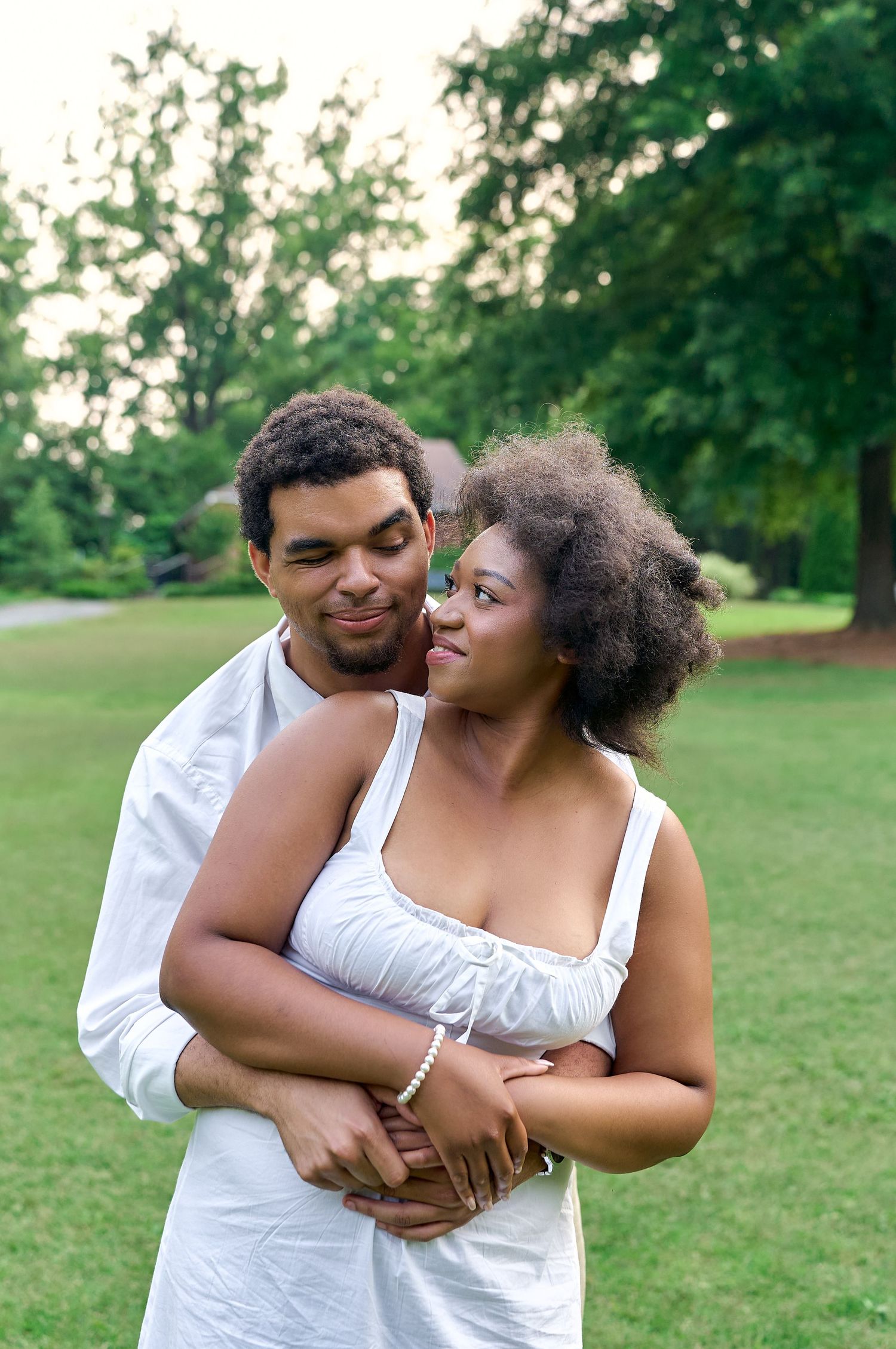 A couple embracing while wearing white outfits against a backdrop of trees in a summer park setting.