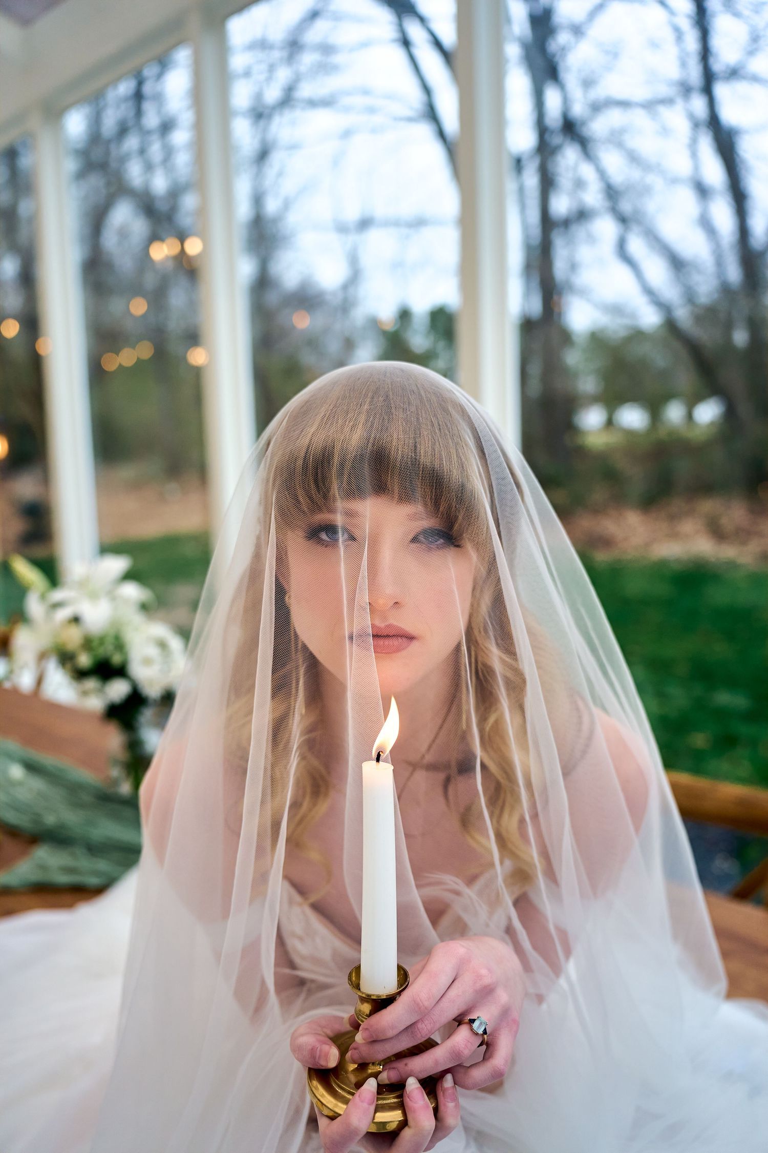 A bride in a white veil holds a lit candle while looking at the camera, with twinkle lights and trees in the background.