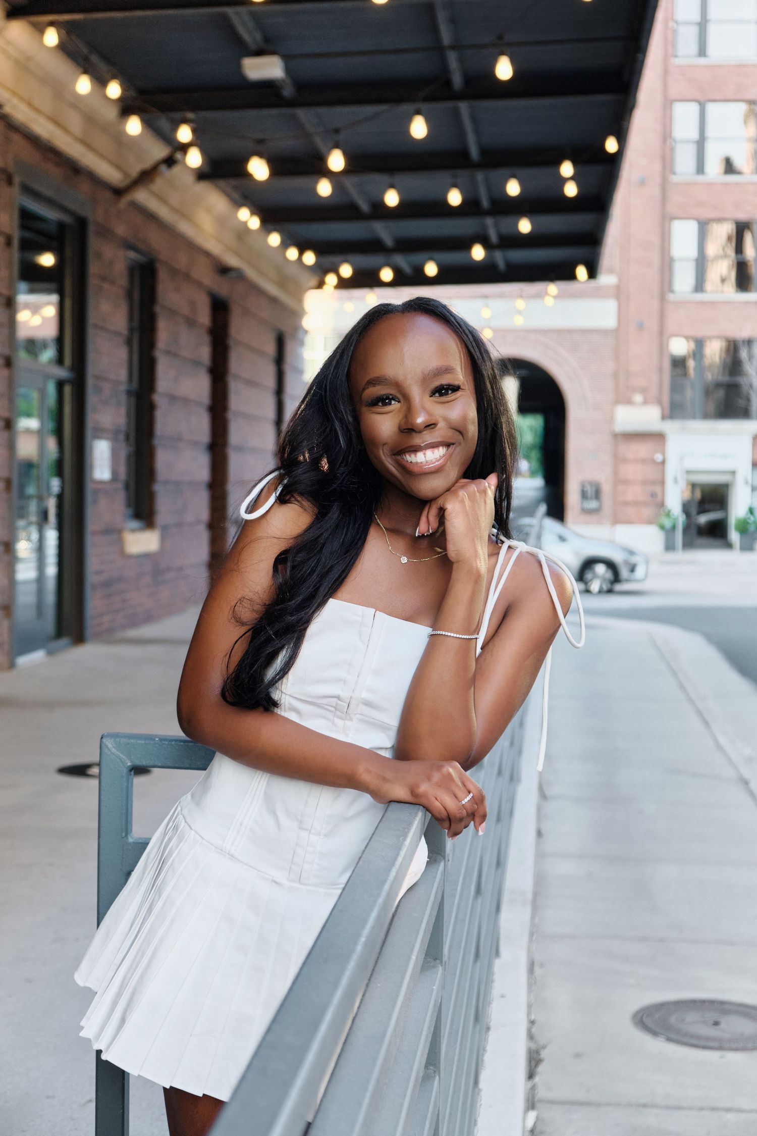 Smiling young woman in white dress leaning on railing with string lights on urban street. North Carolina Photographer.