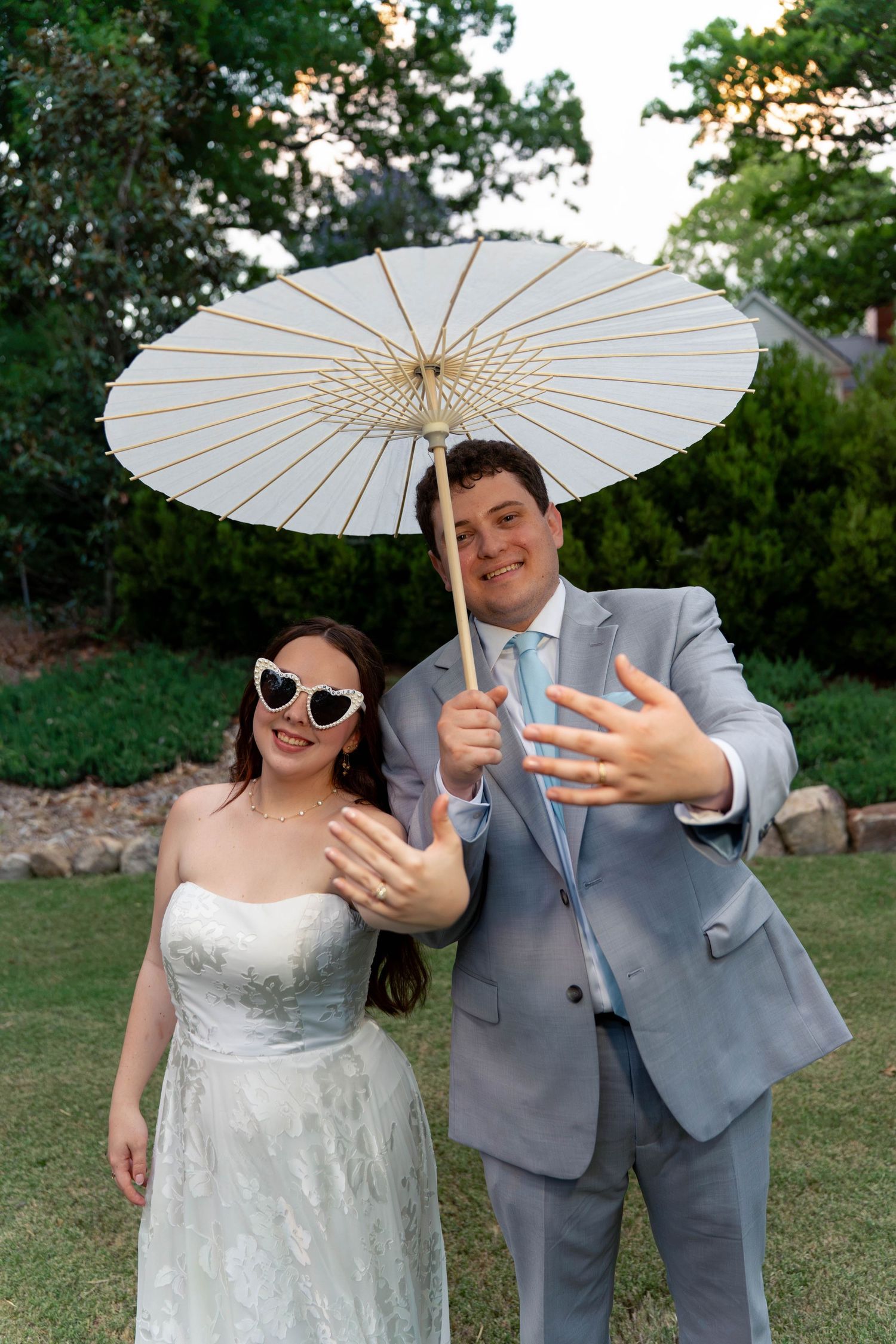 Joyful bride in heart sunglasses and groom under white parasol showing off wedding rings at outdoor reception.