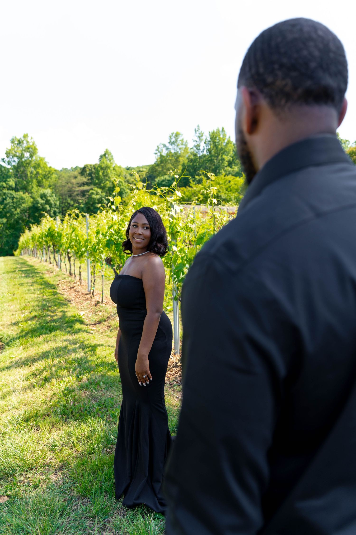 Woman in elegant black gown smiling at partner in vineyard during outdoor engagement photoshoot.