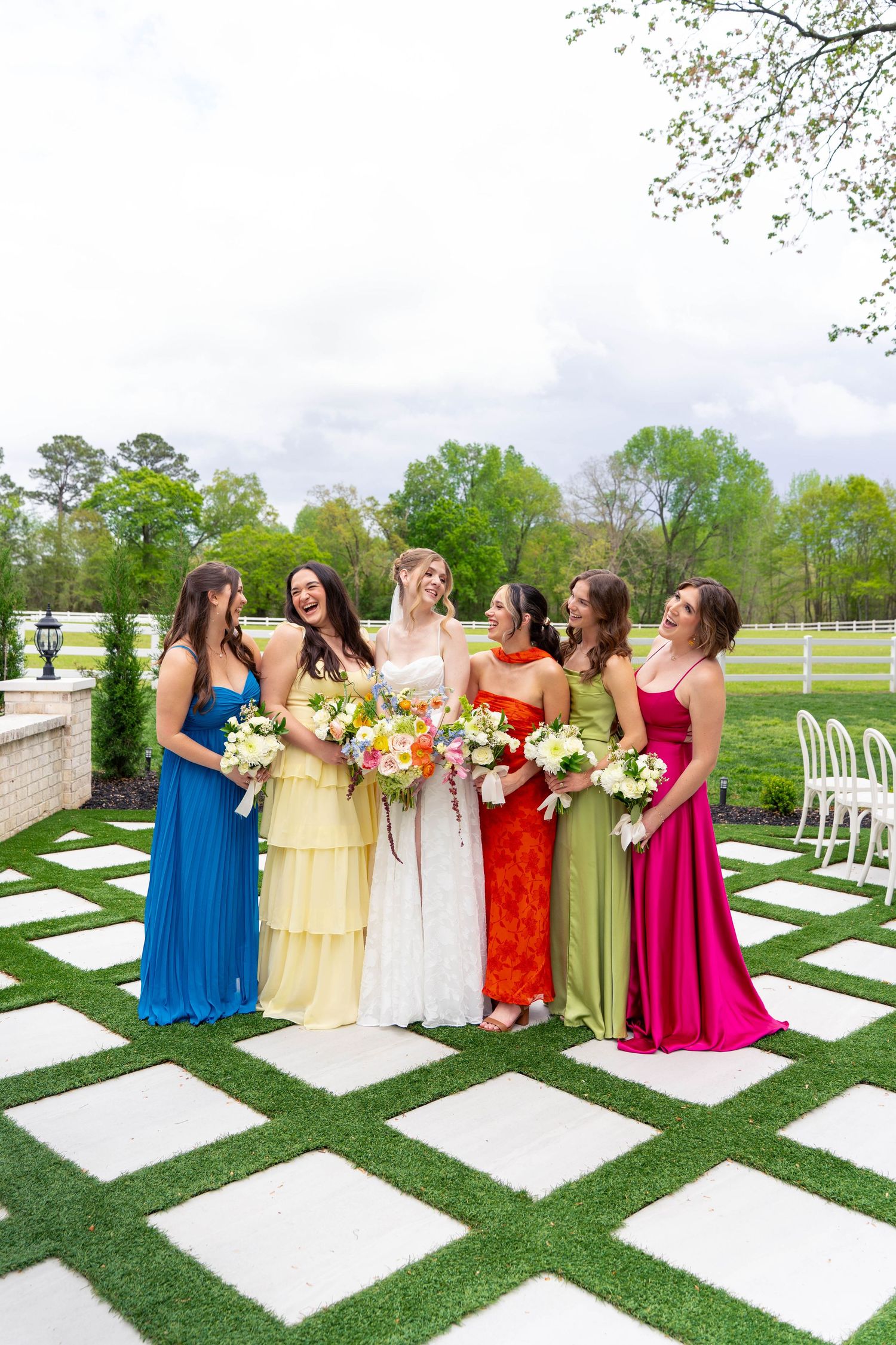 Bride with colorful bouquet laughing with five bridesmaids in rainbow-colored gowns outdoors.