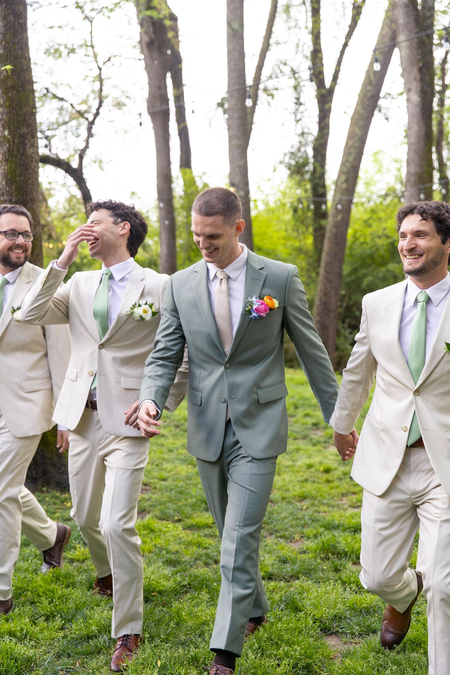 Groom in sage green suit walks with laughing groomsmen in cream suits through a wooded outdoor setting.