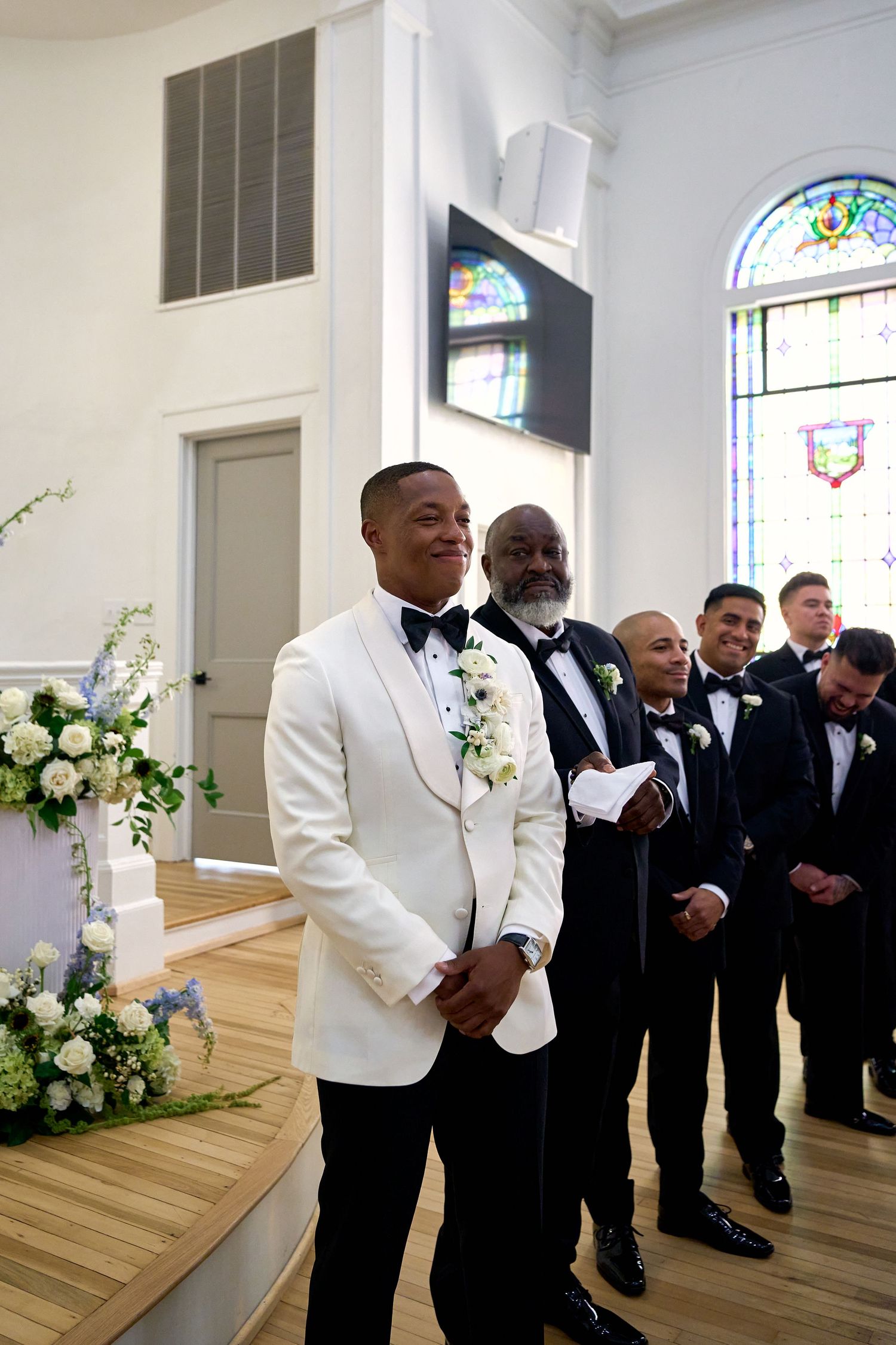 Groom in white tuxedo jacket waits at altar with groomsmen in black tuxedos in church ceremony.