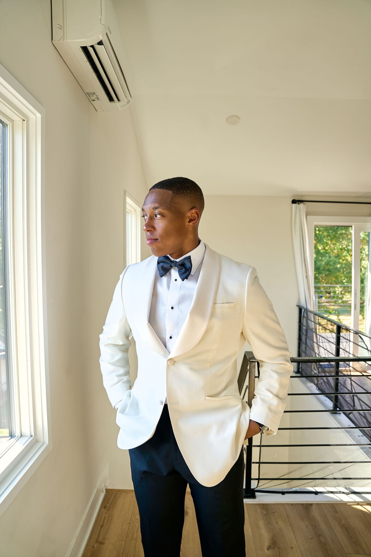 Groom in white dinner jacket and navy bow tie looking out window before wedding ceremony.
