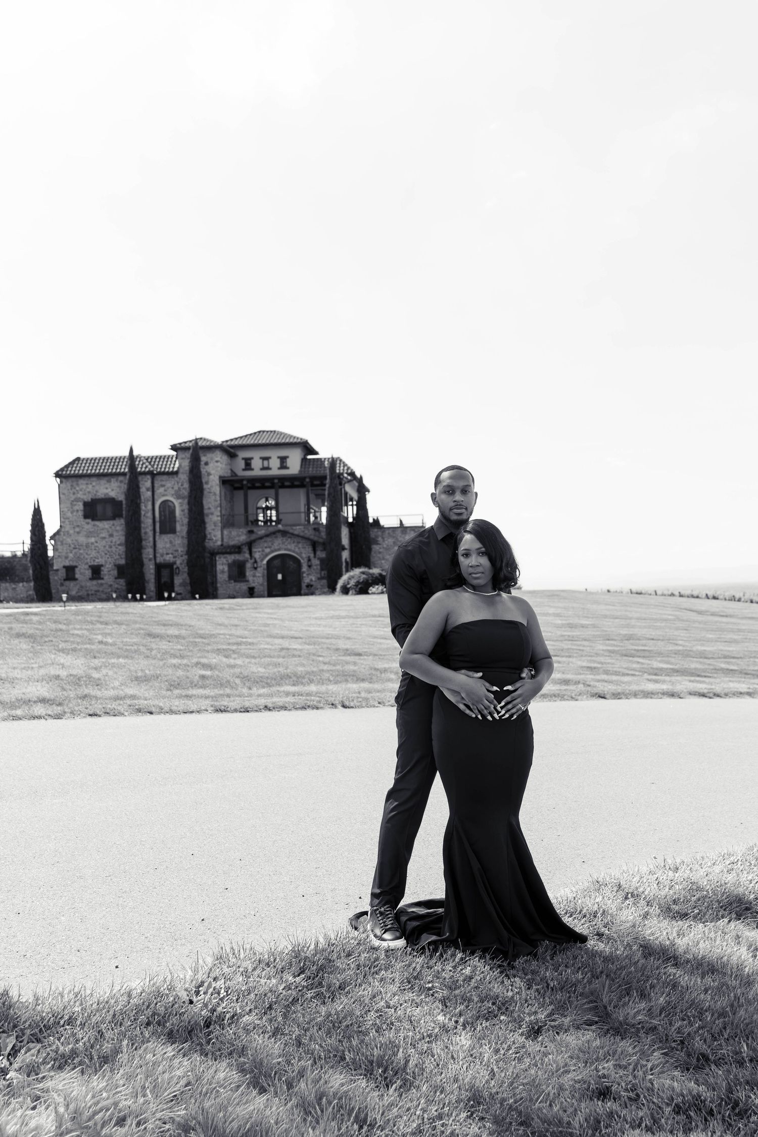 Black and white photo of couple embracing in formal black attire with grand estate in background.