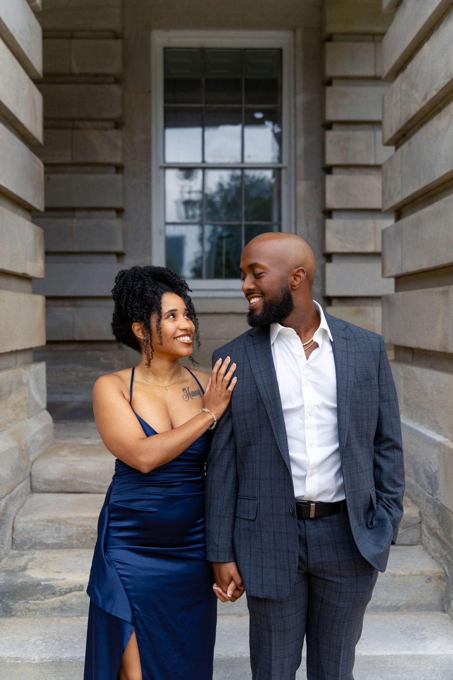 Couple in elegant blue dress and gray suit hold hands and smile on stone steps.