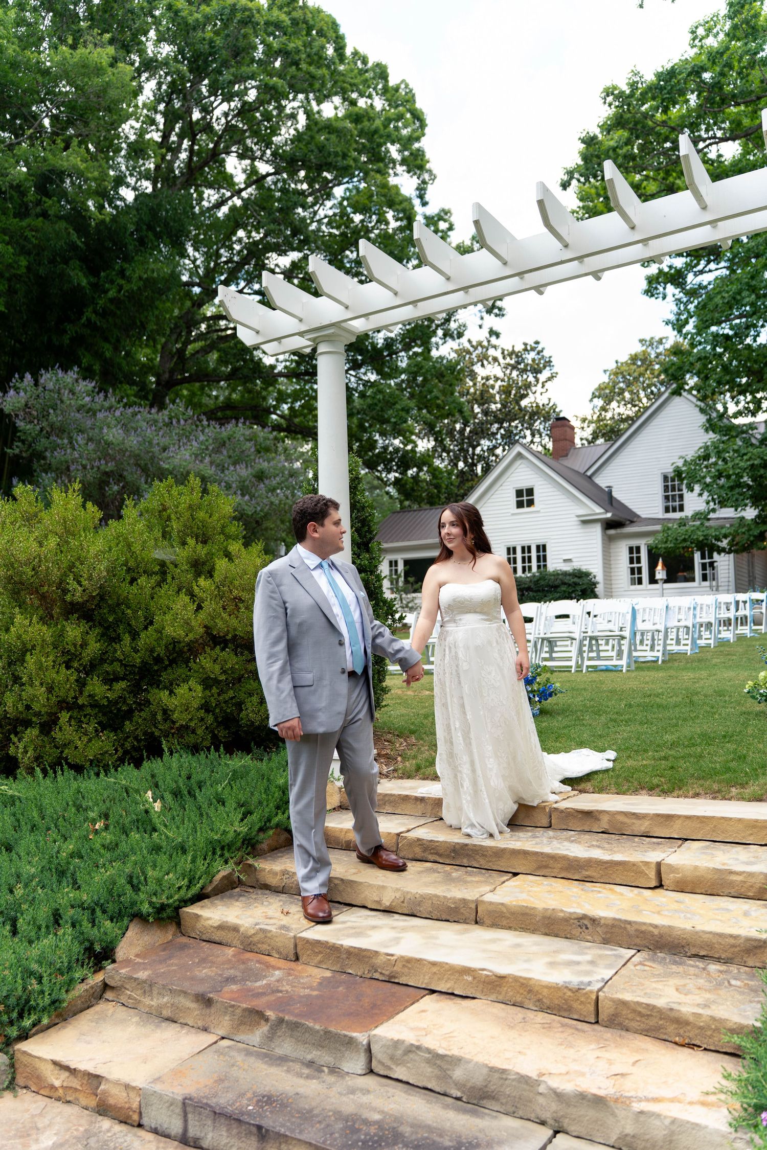 Bride and groom share a romantic moment on stone steps beneath a white pergola in a lush garden wedding venue.