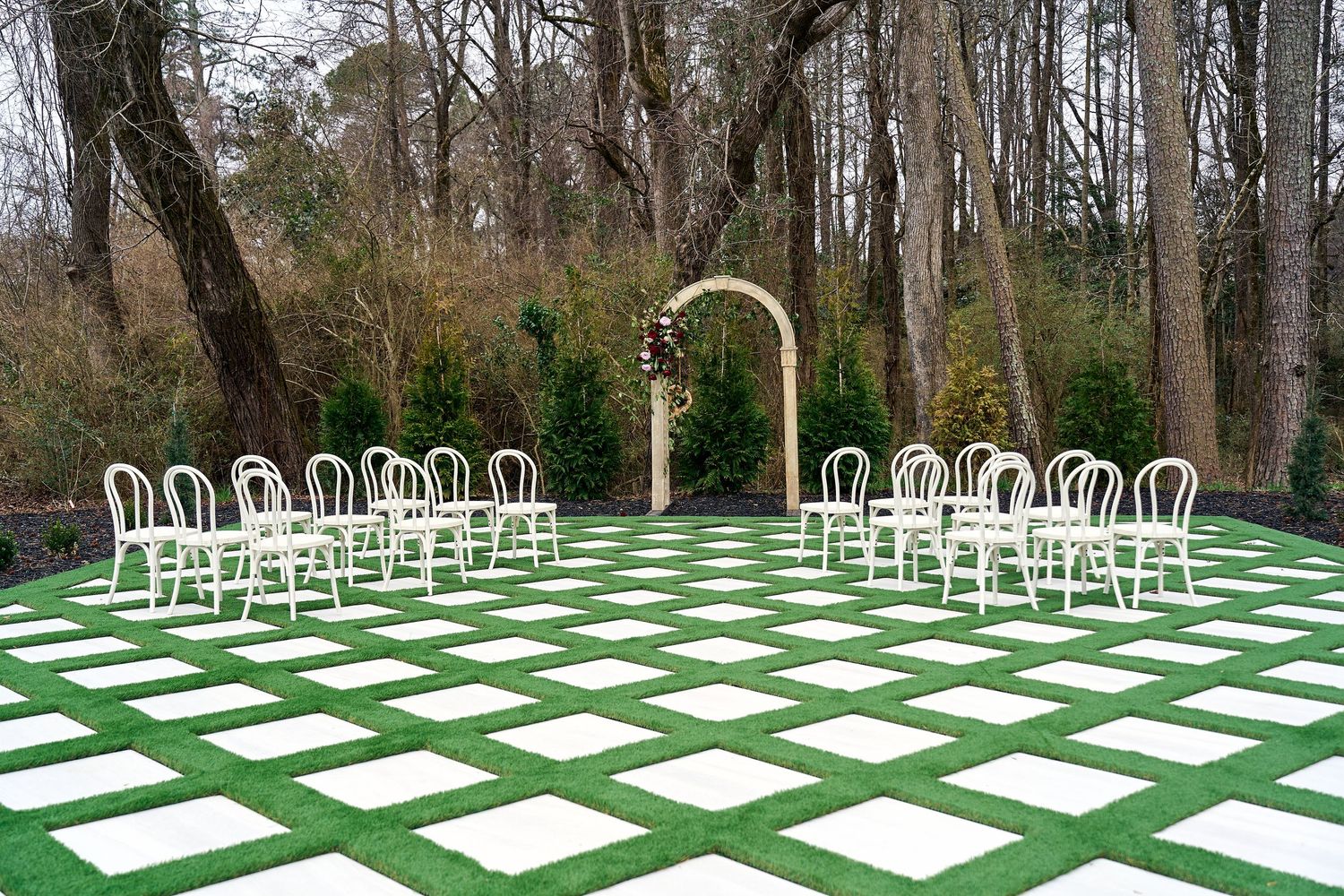 White chairs arranged in rows with a floral arch on a green and white checkered outdoor ceremony space amid trees.